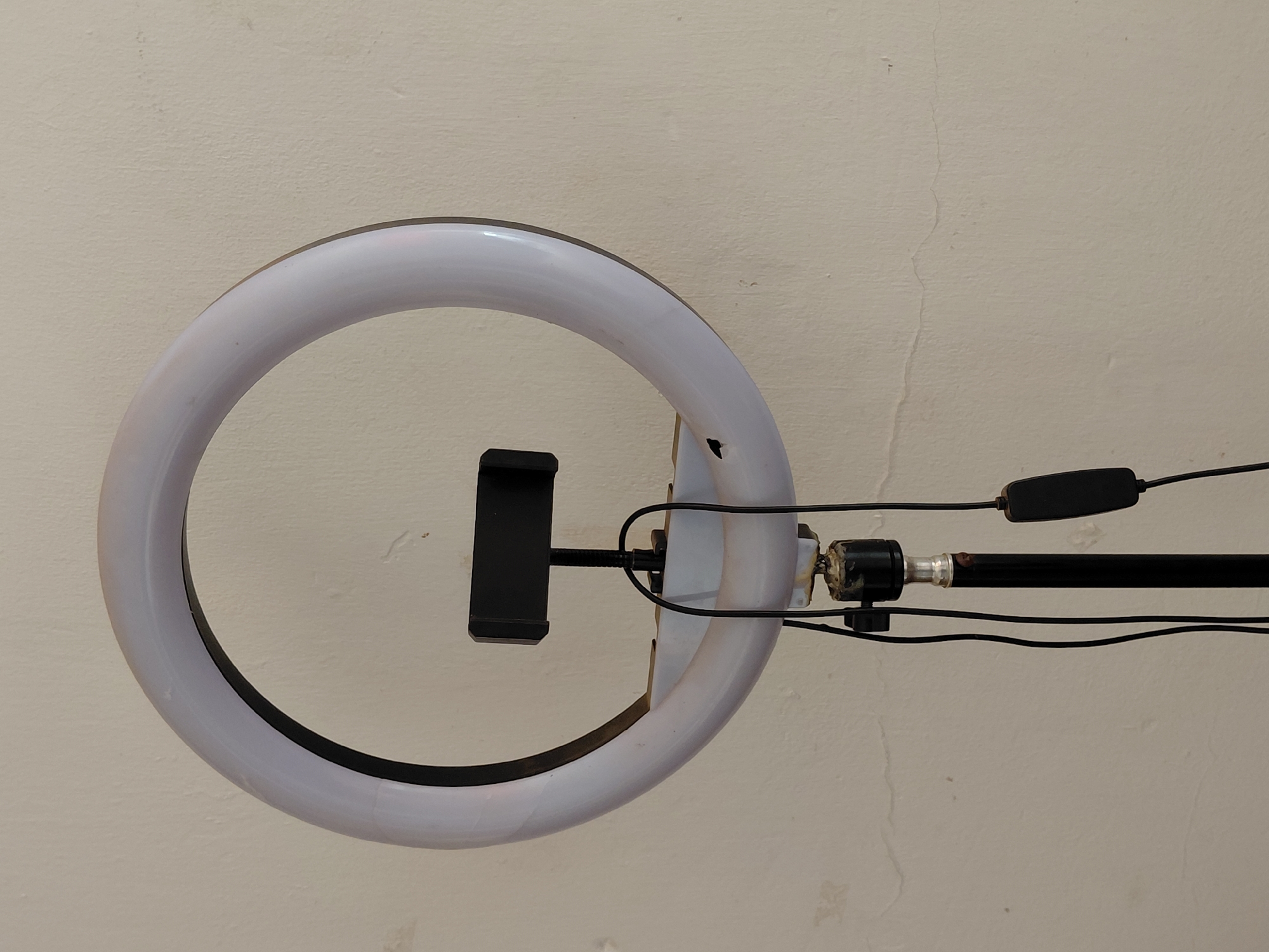 A ring light with an integrated phone holder is positioned prominently against a light-colored, textured wall. The circular ring light features a white, translucent diffuser, suggesting it is illuminated or ready for use. Its inner rim is black, providing contrast. A black, spring-loaded phone holder is centrally mounted within the ring, designed to secure a smartphone for video recording, streaming, or photography.
The ring light is supported on the right by a black metal arm or stand, attached via a swivel joint that shows some signs of wear, including a small amount of adhesive or residue. Black electrical wires emanate from the light, connecting to a small, oblong black control unit visible further along the support arm. These wires are neatly arranged around the stand.
The background is a plain, light cream or beige-colored wall, characterized by a rough, stucco-like texture. Minor imperfections such as faint, vertical cracks and small, scattered dark spots or dust are visible on the wall surface. This suggests an indoor setting, likely in a functional space in Santa Ana, Venezuela, dedicated to content creation. There is no visible text in the image, and the time of day and weather conditions are not discernible.