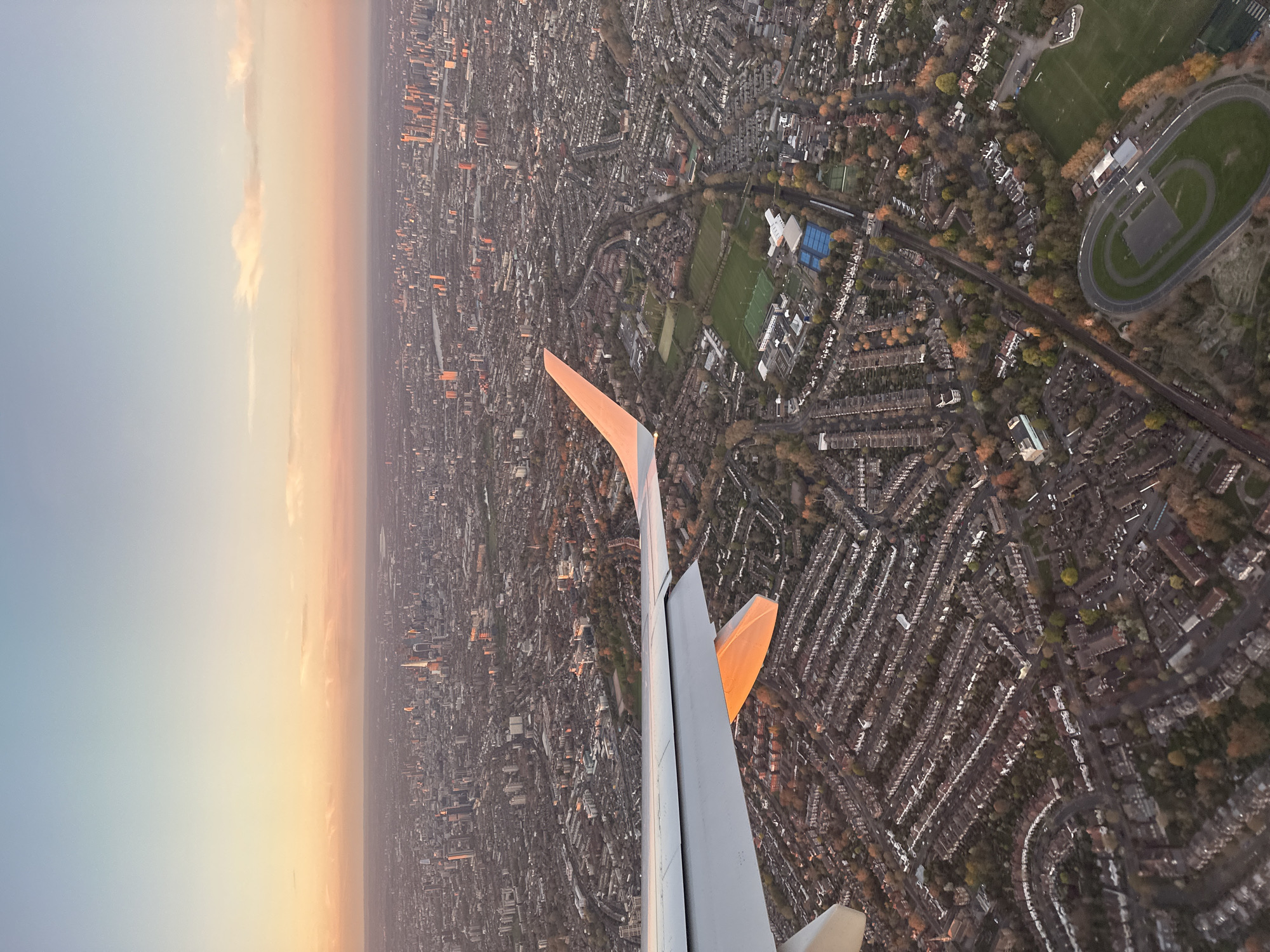 The image captures an aerial view of London, United Kingdom, at dawn or dusk, with the city sprawling beneath the wing of an aircraft. The city is densely populated with rows of tightly packed houses intersected by streets. Green spaces are visible, including playing fields and a running track. The light is soft, creating a warm glow over the city, indicating either sunrise or sunset.