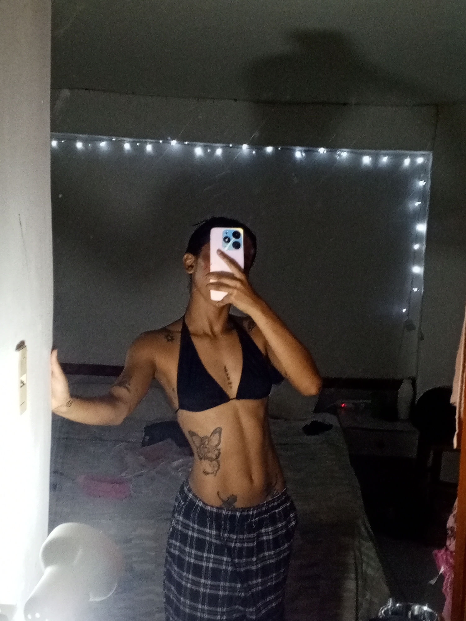 A slender, light-skinned individual is taking a selfie in a bedroom. The person is wearing a black halter top and plaid pajama pants. Tattoos are visible on their arms, stomach, and chest. The person is holding a pink smartphone with a triple-lens camera, obscuring their face. The bedroom has a bed, a nightstand with a light, and string lights hanging along the back wall. The room appears to be dimly lit. The location is likely indoors, but there's no concrete evidence in the image to confirm it being in Porlamar, Venezuela.