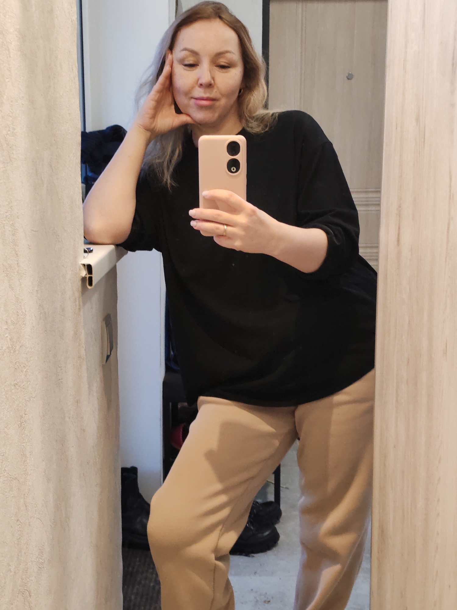 A woman is posing for a selfie in what appears to be an indoor setting, possibly a bathroom or dressing area. She is fair-skinned with blonde hair and is wearing a black long-sleeved shirt and tan sweatpants. She is holding a pink smartphone up to take the photo. Her left hand is resting on a white countertop, and she is looking directly at the camera. A mirror is visible in the background, reflecting the room and the woman herself. In the reflection, black boots can be seen on the floor near the corner of the room. The walls are textured and light in color, and there is a light wood-colored door in the background. The location is in the city of Chaikovsky, Russia.