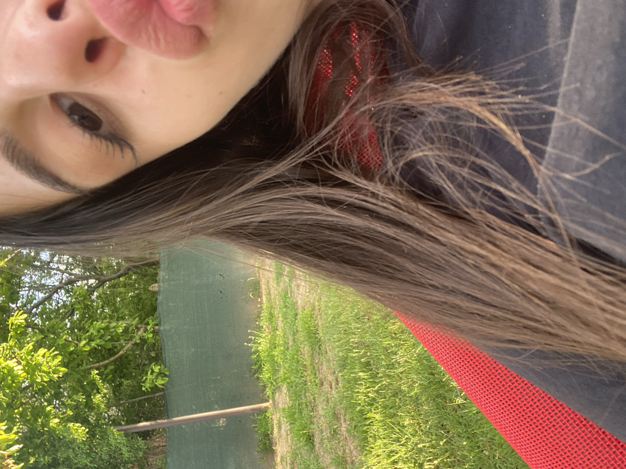 A selfie-style image shows a portion of a person's face, including an eye, eyebrow, nose, and lips. The person has long, dark brown hair with some lighter highlights. A red, mesh-like object is visible near the hair. The background consists of green grass, a fence, and trees. The location is within the city of Unknown, Unknown.