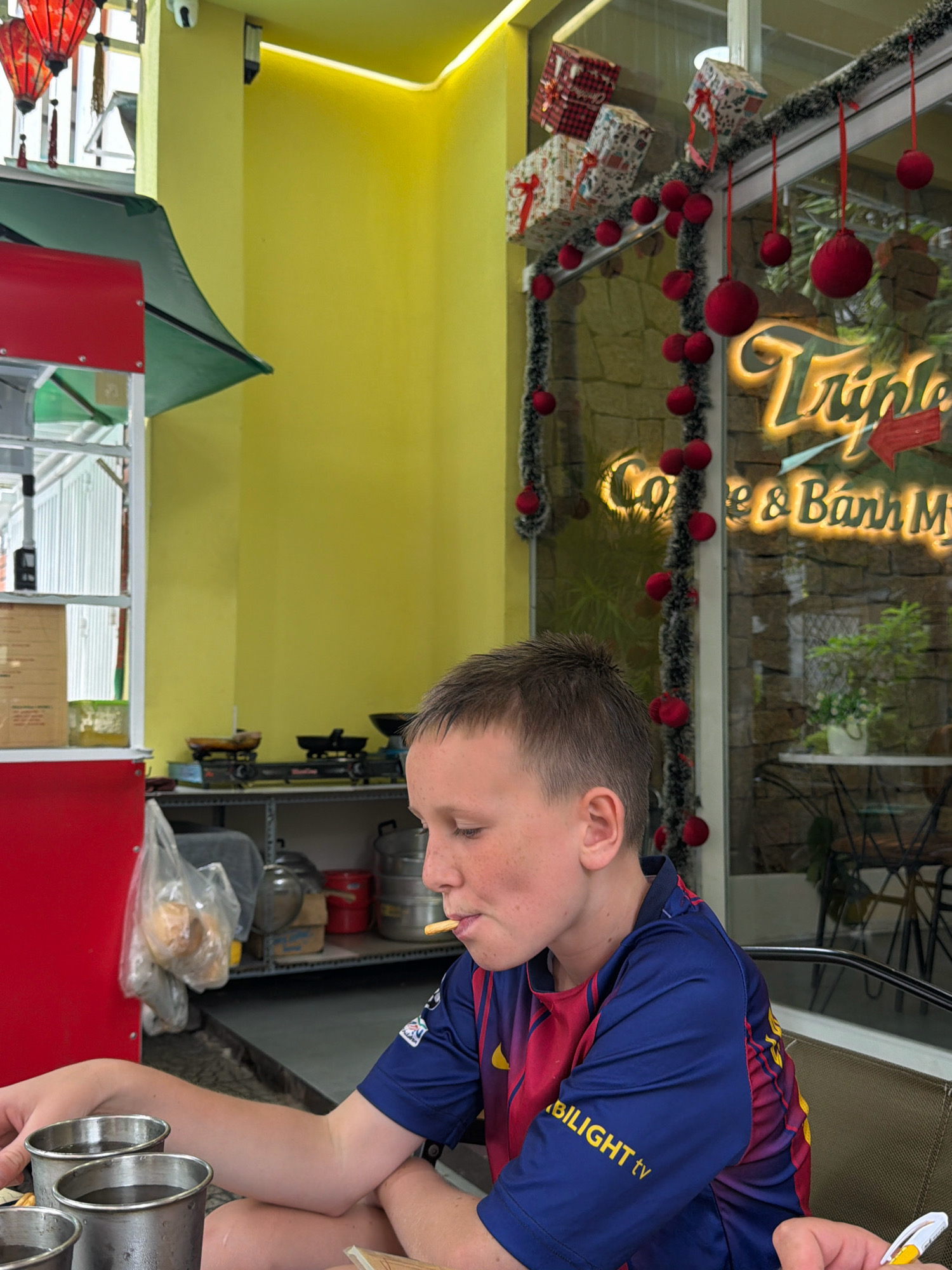 The image depicts a young Caucasian boy with light brown hair, wearing a blue and red soccer jersey, sitting at a table. He is in the process of eating a snack, holding a piece of food in his mouth and reaching for a pen with his right hand. In front of him are three silver cups containing a dark liquid. The background suggests an indoor/outdoor seating area of a restaurant, possibly a cafe, with yellow walls and a stone-patterned wall visible through a glass window. Christmas decorations, including red ornaments and gift boxes, are present along the window. The restaurant's signage, partially visible through the window, reads "Triple Coffee & Banh Mi". The presence of "Banh Mi" in the signage strongly suggests the location is Vietnam, and the style of the restaurant and the general ambiance is consistent with establishments found in cities like Đà Nẵng. A red food cart with a green awning is partially visible on the left side of the image, further indicating a street-food or cafe setting common in Vietnamese cities. The overall atmosphere appears casual and lively.
