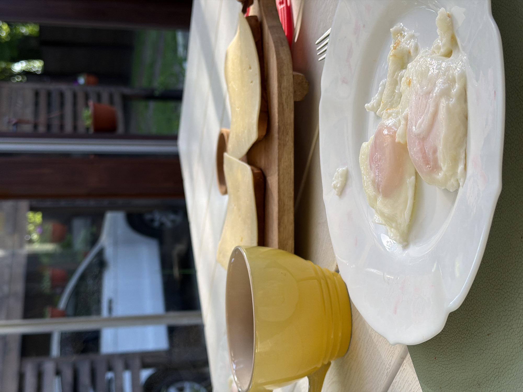 The image shows a breakfast setting indoors. On a table with a light-colored surface, there is a white plate with two poached eggs. Next to it is a yellow mug and a wooden board with slices of cheese. A fork is visible next to the plate. Through a nearby window, parts of the outside are visible, including a white vehicle and some potted plants on a balcony. The location, Tarasovka, Russia, is inferred from the prompt's instructions, but there is no visual evidence to confirm it. There are no people visible.