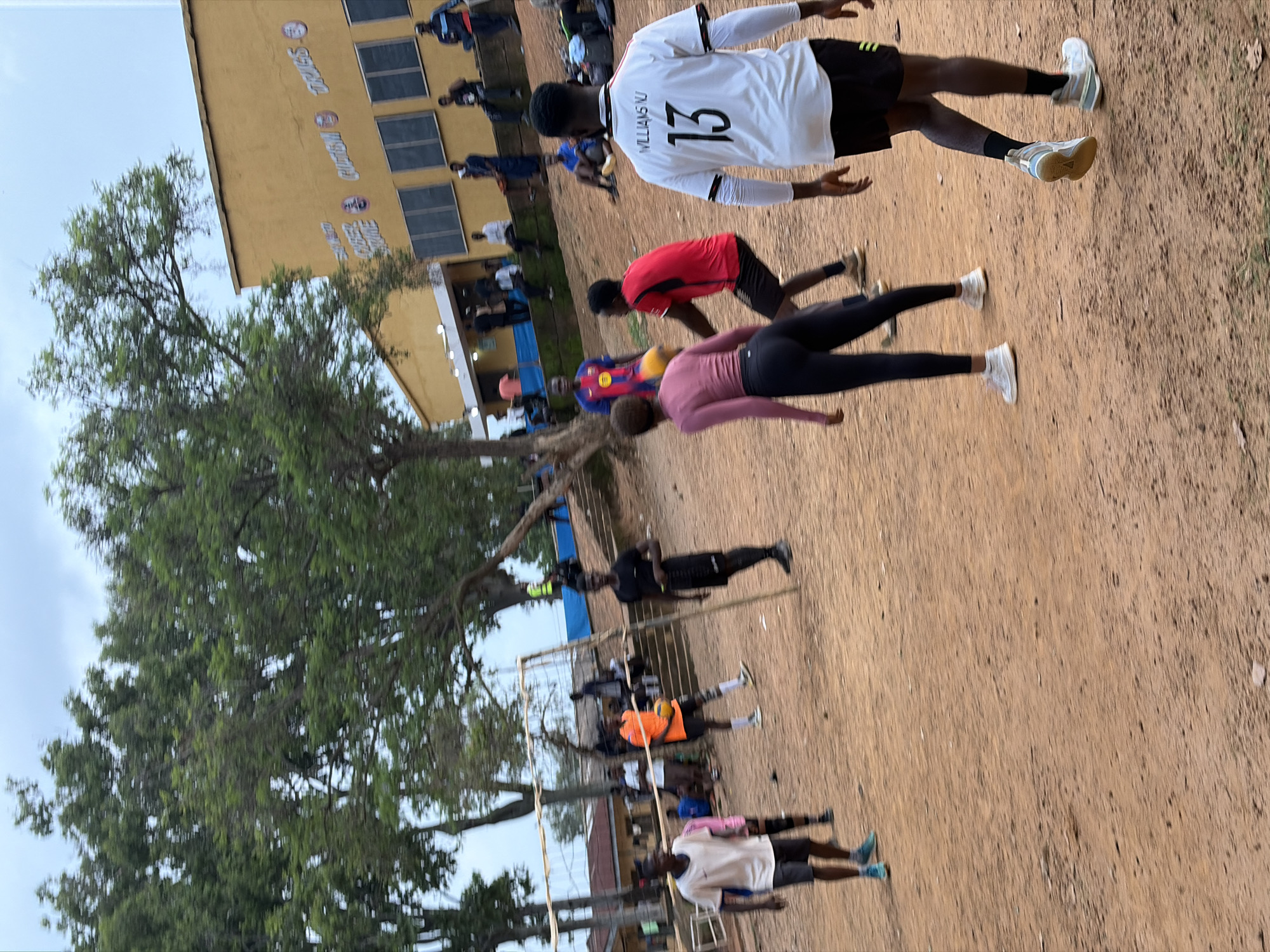 This media file captures a group of people engaged in a physical activity, likely a sports practice or training session, outdoors in Rafin Par, Nigeria. The scene is set on a dirt field in front of a yellow building with multiple windows. A large tree with green foliage is visible to the left, providing some shade. The sky is overcast, suggesting a cloudy day.
The main subjects are individuals in athletic wear, with several in the foreground appearing to be performing exercises or drills. One person in the foreground wears a white jersey with the number "13" and the name "WILLIAMS OU" on the back. Another individual wears a red shirt and black shorts, while a third is in a pink long-sleeved shirt and black leggings. Further back, other individuals can be seen in various colored athletic attire. A volleyball net is set up in the middle ground, suggesting a possible volleyball practice.
The activity appears to be focused on physical conditioning, with individuals in different poses that indicate movement and exertion. The overall atmosphere seems active and focused.
Visible text includes "WILLIAMS OU 13" on the white jersey. The yellow building in the background has some lettering that is partially obscured but appears to be signage related to an institution. The overall impression is that of a sports event or training session taking place in a school or community setting. The time of day is difficult to ascertain precisely due to the overcast conditions, but it appears to be daytime.