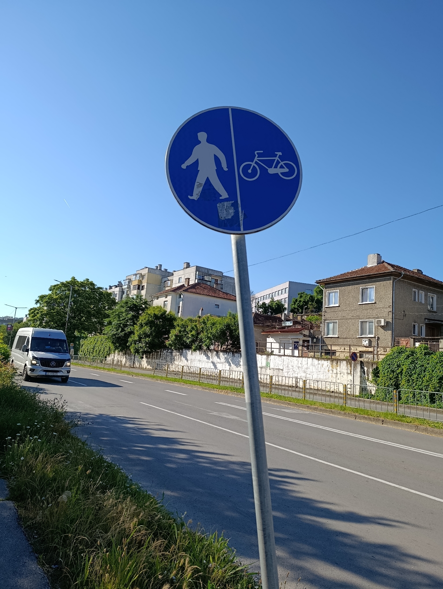 A street sign with a blue circular background and white symbols is positioned on a metal pole in the foreground. The sign is divided vertically, with a pedestrian symbol on the left and a bicycle symbol on the right. A silver van is driving on the asphalt road, which has white lane markings. Tall green trees and residential buildings are visible in the background, under a clear blue sky. The scene appears to be in Razgrad, Bulgaria.