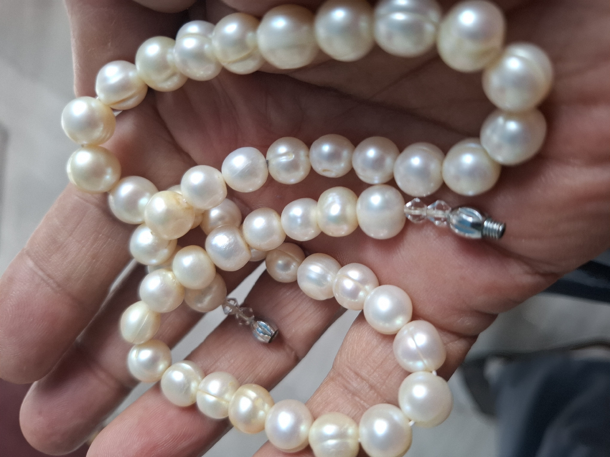 A hand is holding a strand of pearls. The pearls are white and round, with a slight iridescence. They are strung together on a white thread, with a small silver clasp. The background is out of focus, but appears to be a light-colored surface. There is no discernible location in the city of Almaty, Kazakhstan.