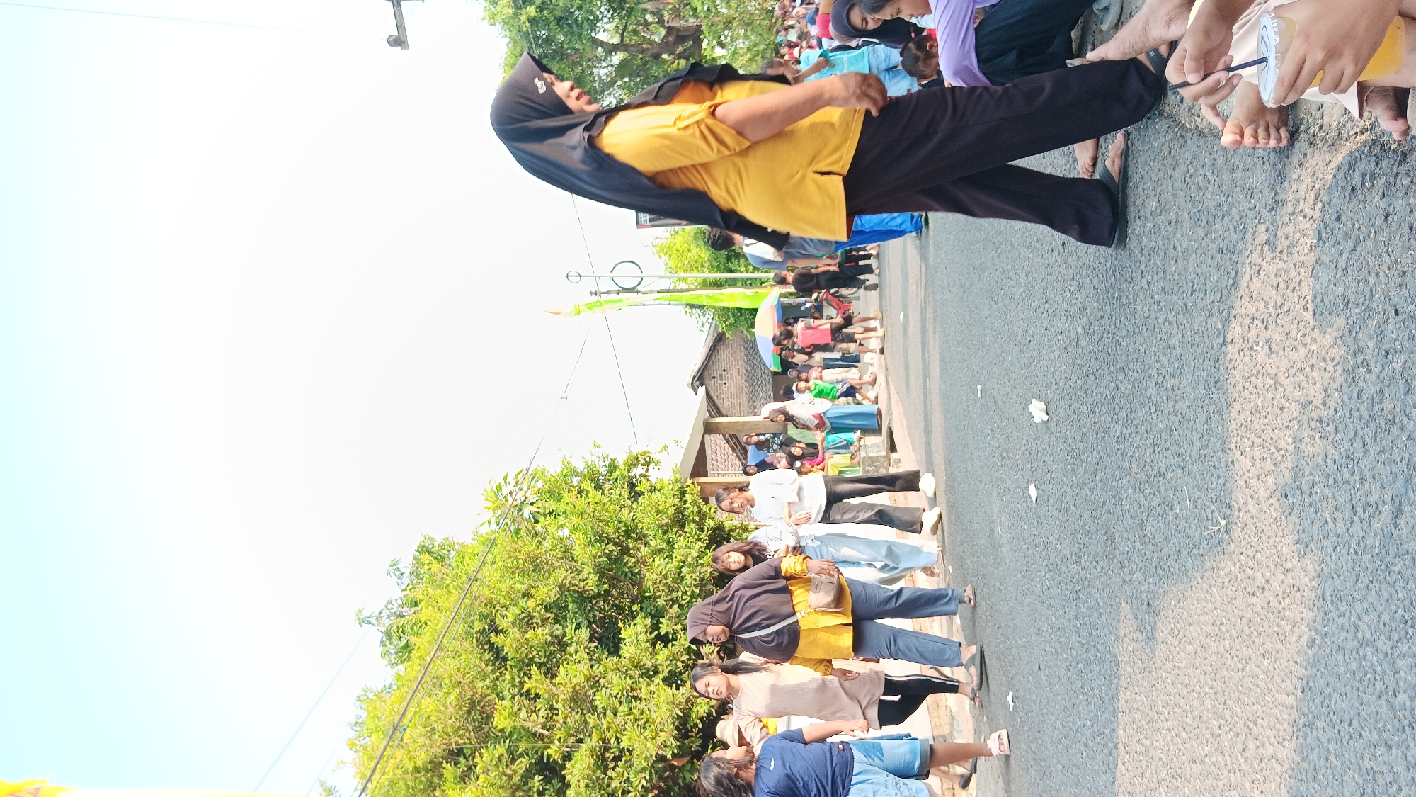 In the city of Cerme, Indonesia, a street scene unfolds under a clear, bright sky. A group of individuals, predominantly women and children, are gathered along the side of a paved road. A woman in a black hijab and a yellow shirt, with dark pants, is prominent in the foreground, walking away from the camera. To her left, a group of women and girls are also walking, some wearing hijabs. The road itself is asphalt and shows signs of wear. Scattered white paper debris is visible on the ground. In the background, houses with simple architecture and a few trees line the street. A colorful umbrella, possibly a vendor's stall, is partially visible amidst the crowd. The overall atmosphere appears to be that of a community gathering or event, possibly a procession or festival.
