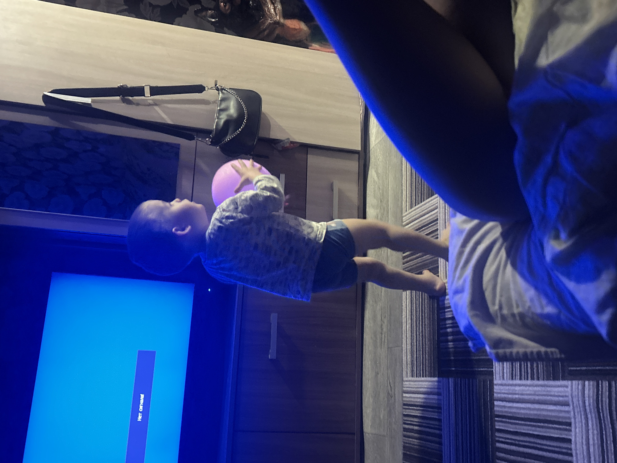 A child is reaching for a pink balloon in a room in Ольховка, Russia. The child is wearing a long-sleeved shirt and shorts. A television with the phrase "Нет сигнала!" is visible. A purse with a strap is on a dresser. A person's legs are visible near the edge of the bed. The floor has a striped pattern.