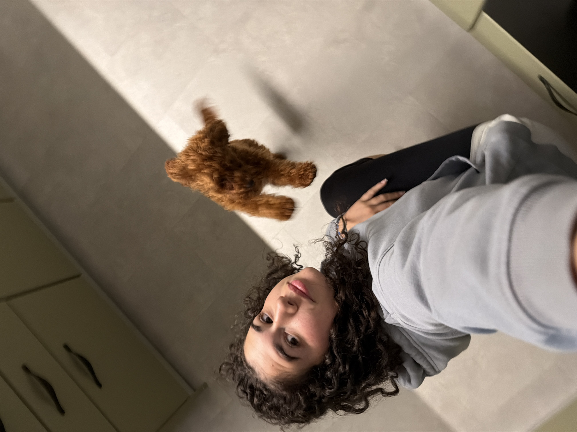 A young woman with dark curly hair is taking a selfie with a small, fluffy, reddish-brown dog in an indoor setting. The woman is wearing a light blue hooded sweatshirt and black leggings. She is sitting on the floor, and the dog, which appears to be a poodle mix, is standing a short distance away, blurred due to motion. The background shows a light-colored tiled floor and cabinets with dark handles. The lighting suggests an interior residential space, possibly a kitchen or hallway. There are no visible indicators of the city of Sofia, Bulgaria.