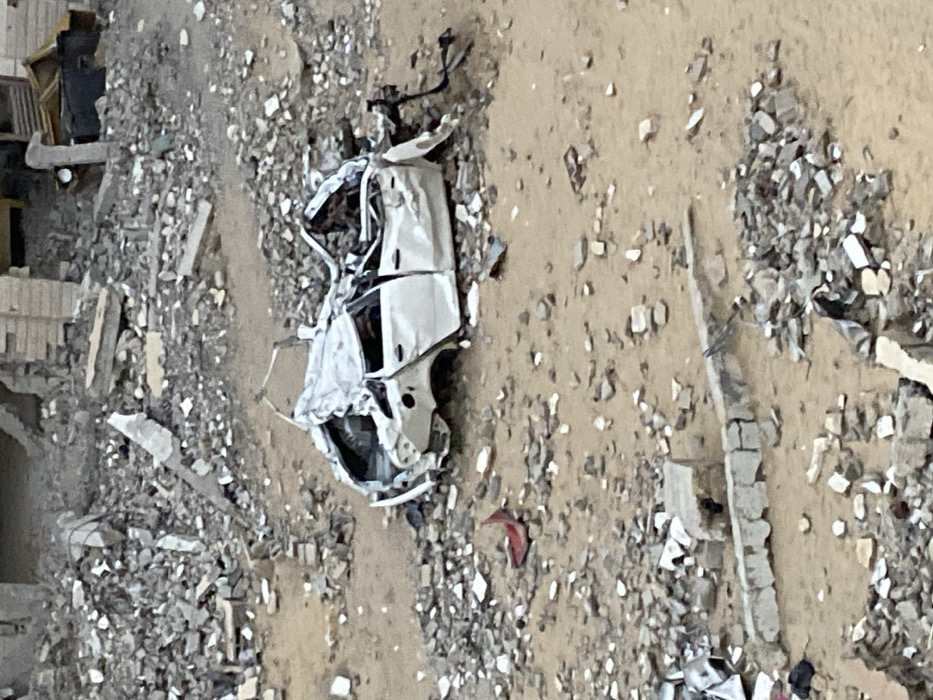 The image depicts a desolate, rubble-strewn urban environment in Khan Yunis, Palestinian Territory. Central to the frame is a severely damaged white four-door sedan, compressed and twisted, with its roof caved in and significant body panels missing or heavily distorted, resting on uneven ground. The surrounding terrain consists of light brown earth mixed extensively with gray and white debris, including concrete fragments, broken bricks, and other building materials. In the upper background, remnants of collapsed structures are visible, composed of piles of similar rubble and fractured sections of walls or foundations. A small red piece of debris lies to the left of the vehicle, and a partially intact concrete barrier runs across the lower portion of the foreground. No individuals or discernible actions are present in the scene.