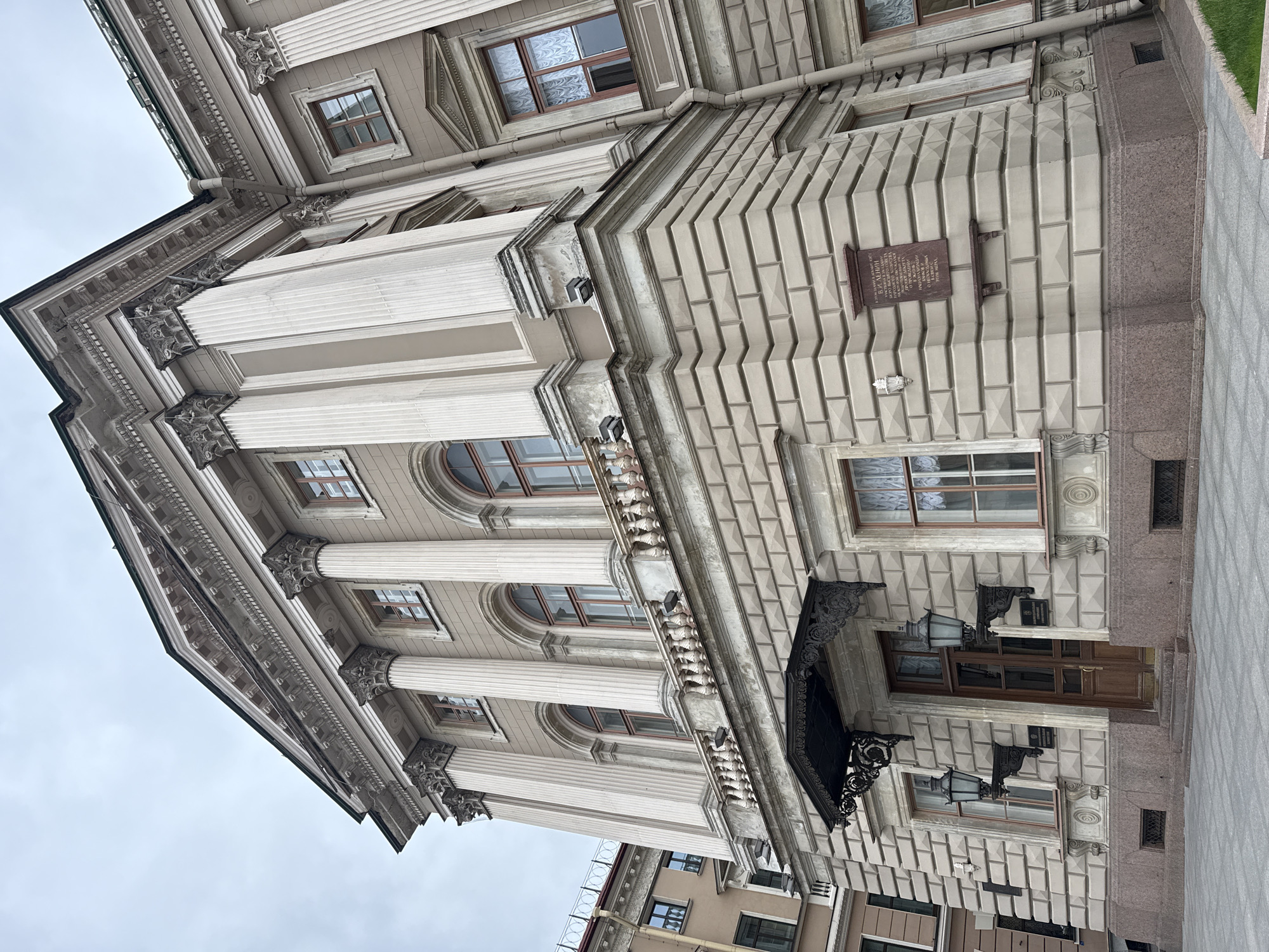 The image showcases a building facade in Saint Petersburg, Russia. The architecture is neoclassical, featuring columns with ornate capitals, arched windows, and detailed stonework. A plaque is visible on the wall, likely containing historical information about the building. An entrance with a canopy and decorative lamps adorns the building's base. The color palette is dominated by shades of beige and gray, reflecting the typical Petersburg architecture. The sky appears overcast.