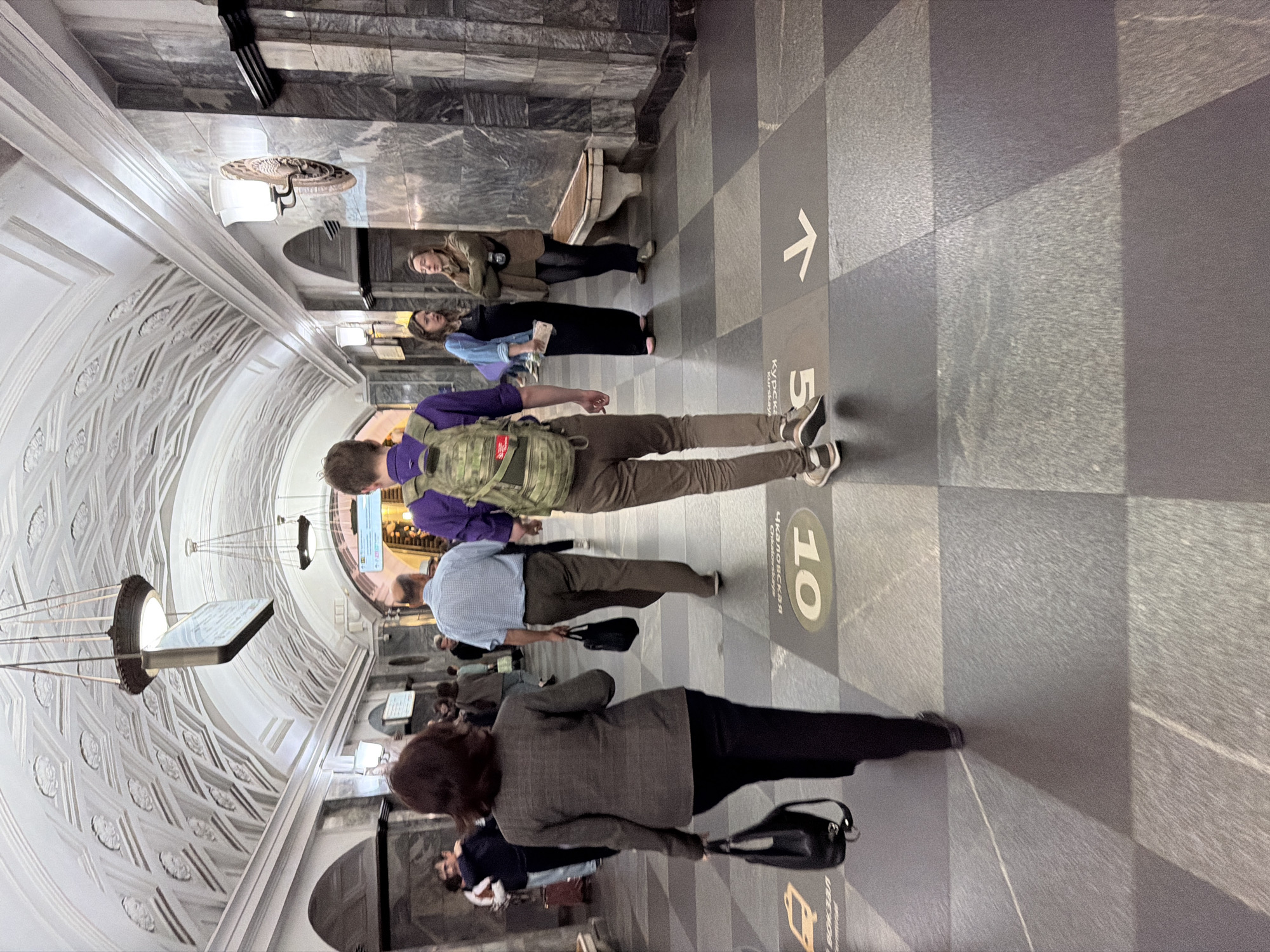 The image shows a wide, ornate subway station platform in the city of Unknown. The ceiling is a vaulted, decorative plaster work with repeating patterns, illuminated by pendant lights. The walls are lined with dark, polished stone panels. In the foreground, a woman with dark hair, wearing a gray blazer and black pants, walks away from the camera, carrying a black handbag. To her right, a man in a light blue shirt and khaki pants also walks away from the camera, carrying a black bag. Further ahead, a young man with a backpack on, wearing a purple shirt and brown pants, is walking towards the camera. In the midground, two women are standing near a bench; one in a black dress and blue jacket, and the other in a brown jacket. The floor is tiled in a checkered pattern of dark gray and lighter gray squares, with directional arrows and station numbers visible on some tiles. Several people are scattered throughout the platform, some walking and some standing.