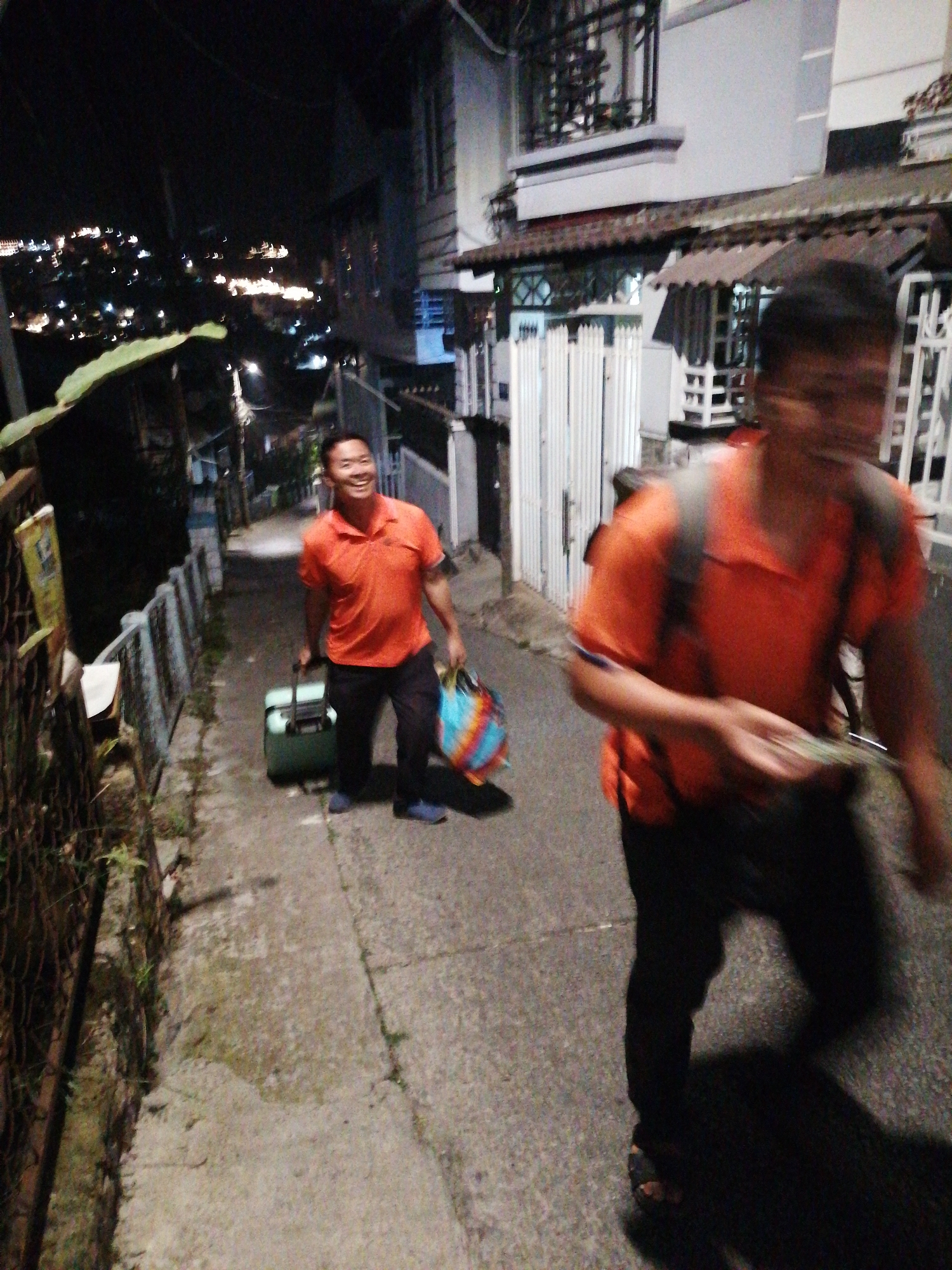 A medium-shot photograph shows two men walking on a narrow, upward-sloping concrete path at night in Da Lat, Vietnam. The man on the left, in an orange polo shirt and black pants, is smiling and pulling a green suitcase with his right hand. He is also holding a multi-colored bag in his left hand. The man on the right is blurred and wearing an orange shirt and dark pants, with a backpack. The path is lined with a metal fence and dense foliage on the left, and buildings with windows and awnings on the right. In the background, the lights of the city spread out across the hillside, indicating a nighttime urban scene. The overall impression is of a street in a hilly area of Da Lat.