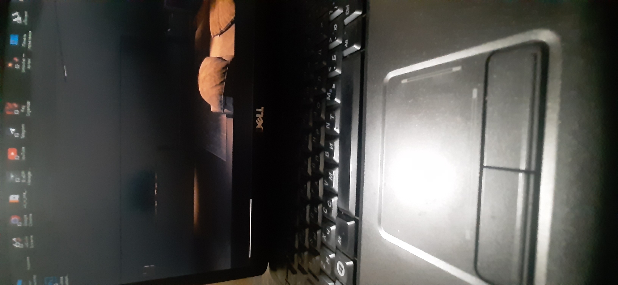 The image displays a close-up, slightly angled view of an open laptop. The laptop lid is down, showing a dark screen with a low-resolution wallpaper depicting what appears to be a room with cushions or pillows. The "DELL" logo is visible on the laptop's bezel. Below the screen is the keyboard, which is partially visible, and the touchpad. The keyboard appears to have Cyrillic characters on the keys. The scene is indoors, with the lighting suggesting it is not a brightly lit environment. There are no people present in the image. The image itself is of moderate quality with some blurriness.
