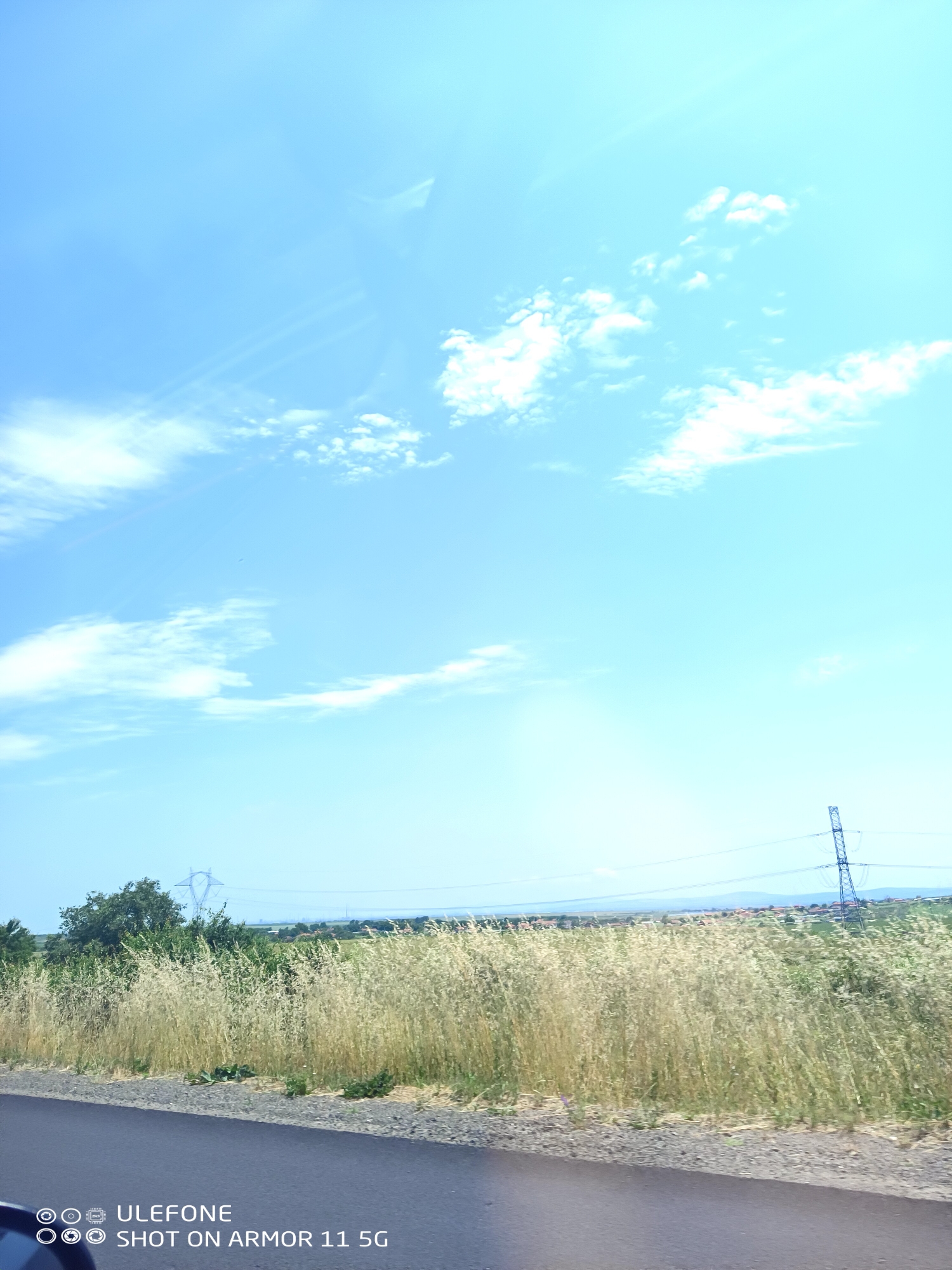 A wide-angle landscape photograph captured in Krustina, Bulgaria, depicts a sunny rural scene dominated by a bright blue sky with scattered white clouds. The sun's intense light generates visible lens flare in the upper sky, particularly in the upper right quadrant and center-left. The foreground features a dark asphalt road with a narrow gravel shoulder, leading into a field of tall, golden-brown dry grass that occupies the mid-ground. Behind the dry grass on the left, denser green foliage, possibly trees or shrubs, is visible. The horizon reveals faint, hazy mountains or hills. Several electricity transmission towers (pylons) are present: one partially visible on the far right, and another, lighter-colored, positioned further back in the center-left. Power lines extend horizontally between these structures. In the distant mid-ground, below the mountains, subtle indications of human habitation, such as buildings, can be observed. No individuals or animals are present. The image, bearing a "Ulefone SHOT ON ARMOR 11 5G" watermark, appears to have been taken from a low vantage point, likely from a vehicle.