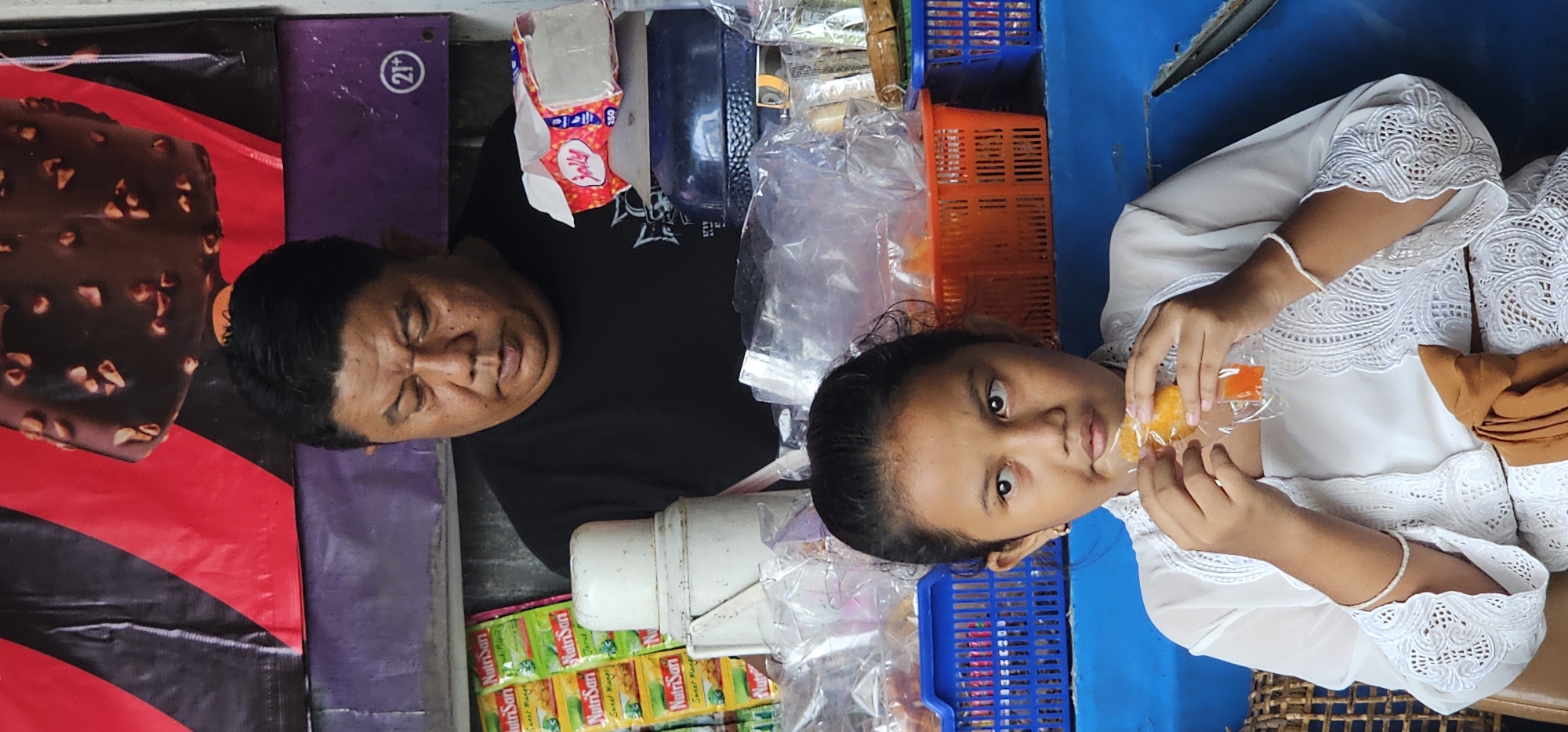 The image appears to be taken at a small street vendor stall in Denpasar, Indonesia. A middle-aged man in a black t-shirt is standing behind the stall. A young girl in a white lace-trimmed top is standing on the opposite side of the stall, holding an unwrapped orange-colored sweet treat. The stall is stocked with various goods, including ice cream advertisements, tissue packs, canned drinks, and packaged snacks. Several plastic crates of assorted colors are visible. The background suggests an urban environment.