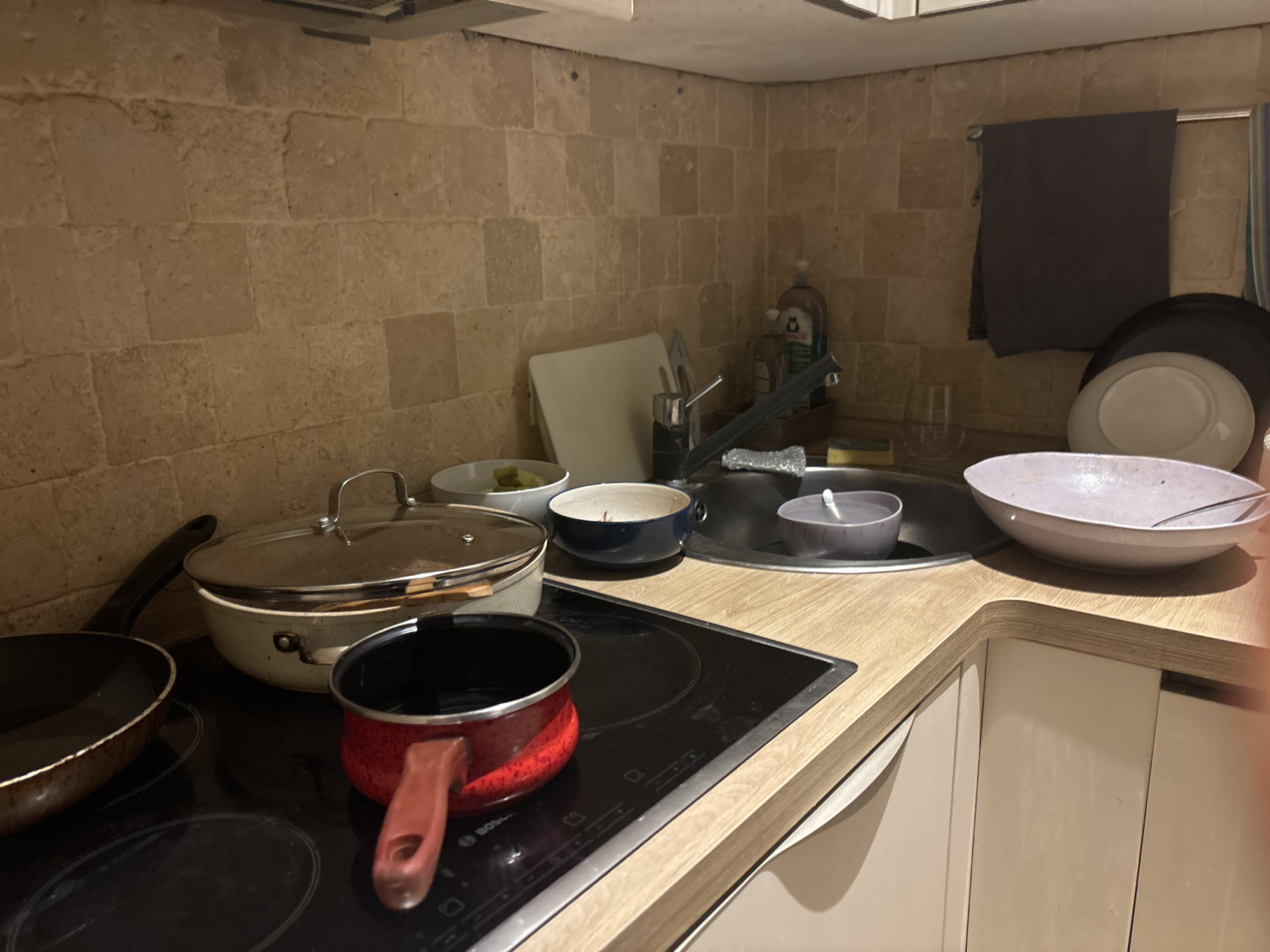 The image shows a cluttered kitchen in what appears to be an apartment. The countertops are overflowing with dirty dishes including pots, pans, bowls, and plates. Some items are in the sink, which also appears full. There is a stovetop with cookware on it, and a dish towel hanging nearby. The kitchen has tiled walls and a countertop with cabinets underneath.
It's not possible to confirm the location of the kitchen as being in Budva, Montenegro, based on the visual information present. There are no identifying geographical features or landmarks visible.