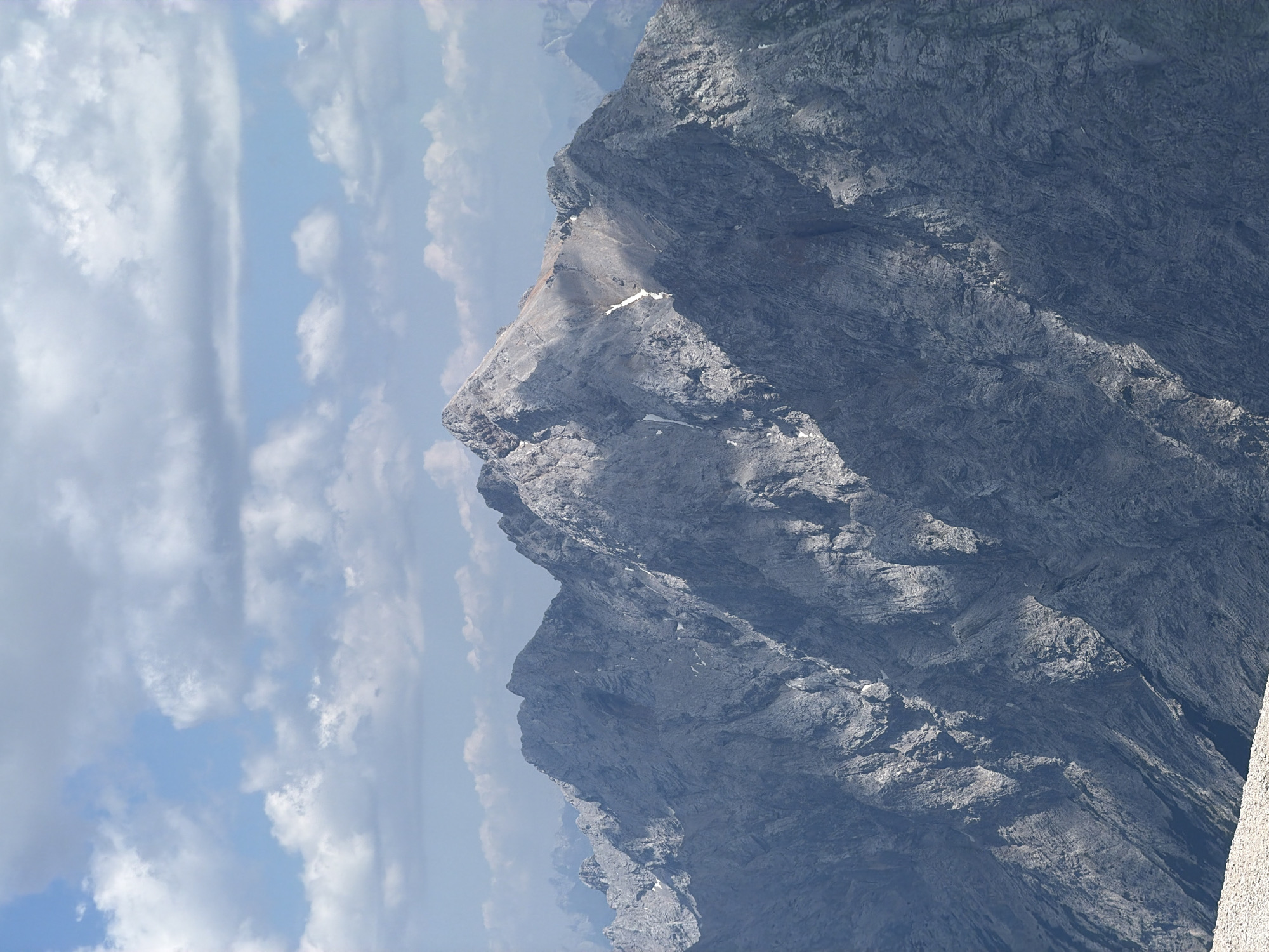 This is a vertical, close-up photograph of a rugged mountain face under a bright, partly cloudy sky. The primary subject is the dramatic, rocky peak, which is characterized by steep slopes, jagged edges, and various textures of grey stone. Sunlight casts sharp shadows, highlighting the contours and creating a sense of depth and scale. Patches of white, likely snow or ice, are visible on the mountain, particularly in crevasses and along certain ledges.
The background consists of a vast expanse of sky, filled with a mix of white, puffy cumulus clouds and streaks of more wispy cirrus clouds. The blue sky peeks through in areas. The overall impression is one of natural grandeur and a remote, high-altitude environment.
There are no people or man-made objects visible in the frame, emphasizing the wild and untouched nature of the scene. The lighting suggests it is daytime, likely late morning or early afternoon, given the strength of the sun and the sharp shadows. The weather appears fair, with clear skies punctuated by clouds. The location context of Garmisch-Partenkirchen, Germany, suggests this is likely a view of the Bavarian Alps, known for their dramatic peaks and challenging terrain. There is no visible text in the image.