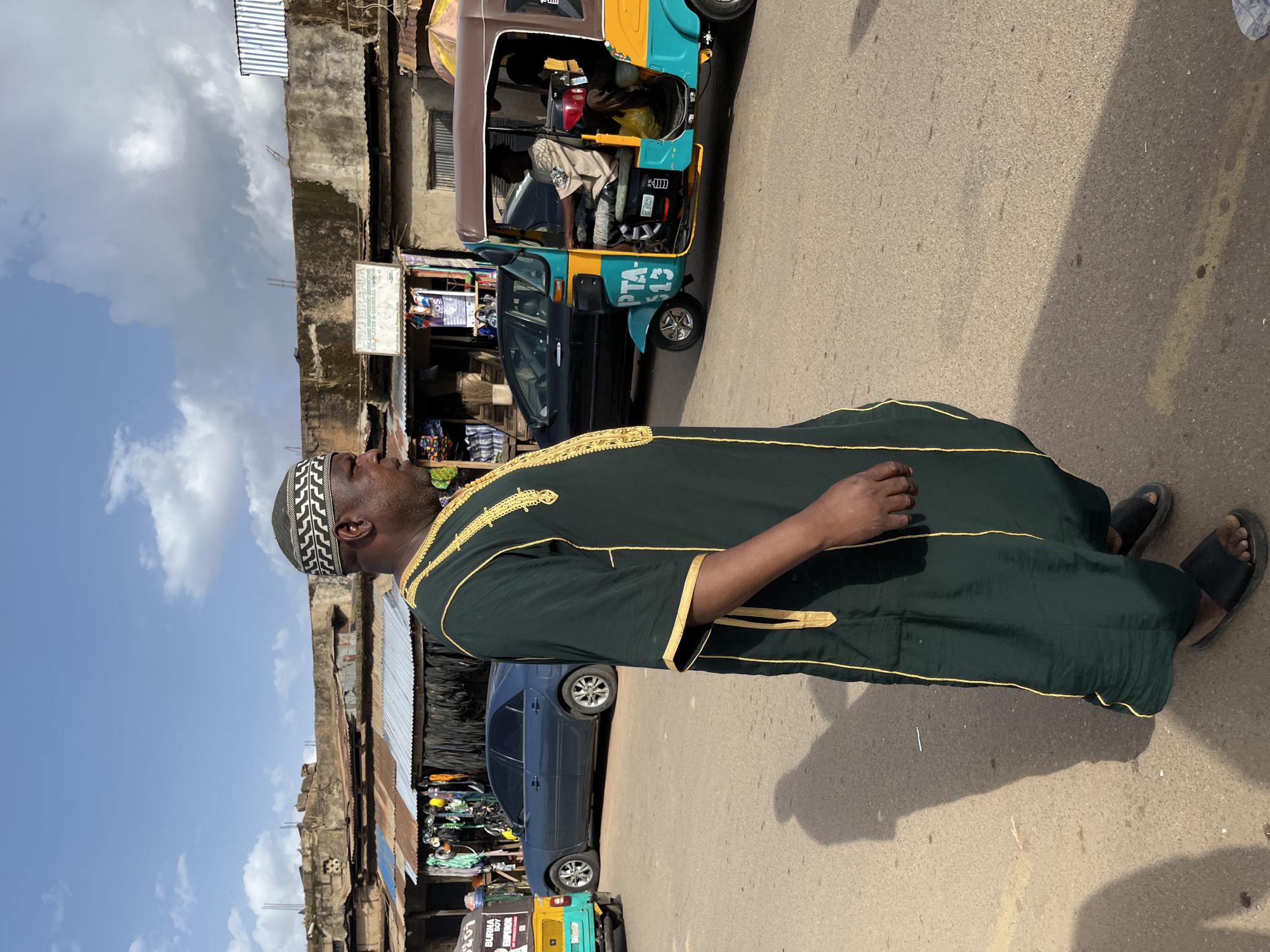 A man wearing a green robe with gold trim and a patterned kufi cap stands on a paved street. He is facing left, with his head turned slightly upwards and to the left. The man is wearing black sandals. Behind him, a colorful auto-rickshaw, painted teal, yellow, and brown, is parked on the street. The auto-rickshaw has text on its side, including "PTA 513". Further behind the auto-rickshaw, a dark blue sedan is visible. The background shows buildings with corrugated metal roofs and weathered walls, suggesting a market or urban setting. The sky is blue with scattered white clouds, indicating it is daytime and the weather is fair. The scene appears to be in a busy area with signs of commercial activity.