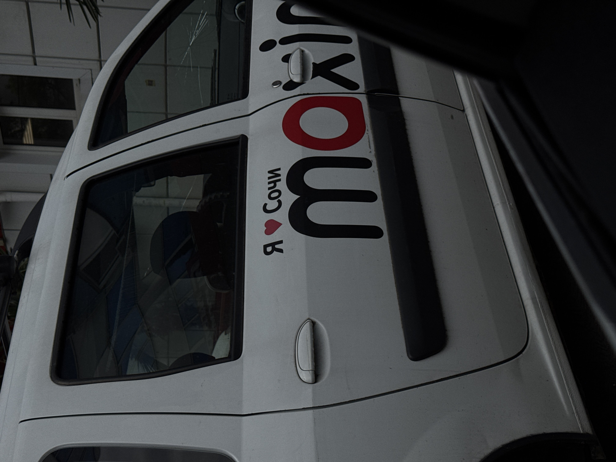 A white car with the word "Maxim" displayed vertically on its side is partially visible. Below the word, in Russian, is "I love Sochi" with a red heart symbol. The car has a cracked windshield. It appears to be parked in Sochi, Russia. The background shows a building with windows, and part of a palm tree is visible. The image is taken from inside another vehicle.