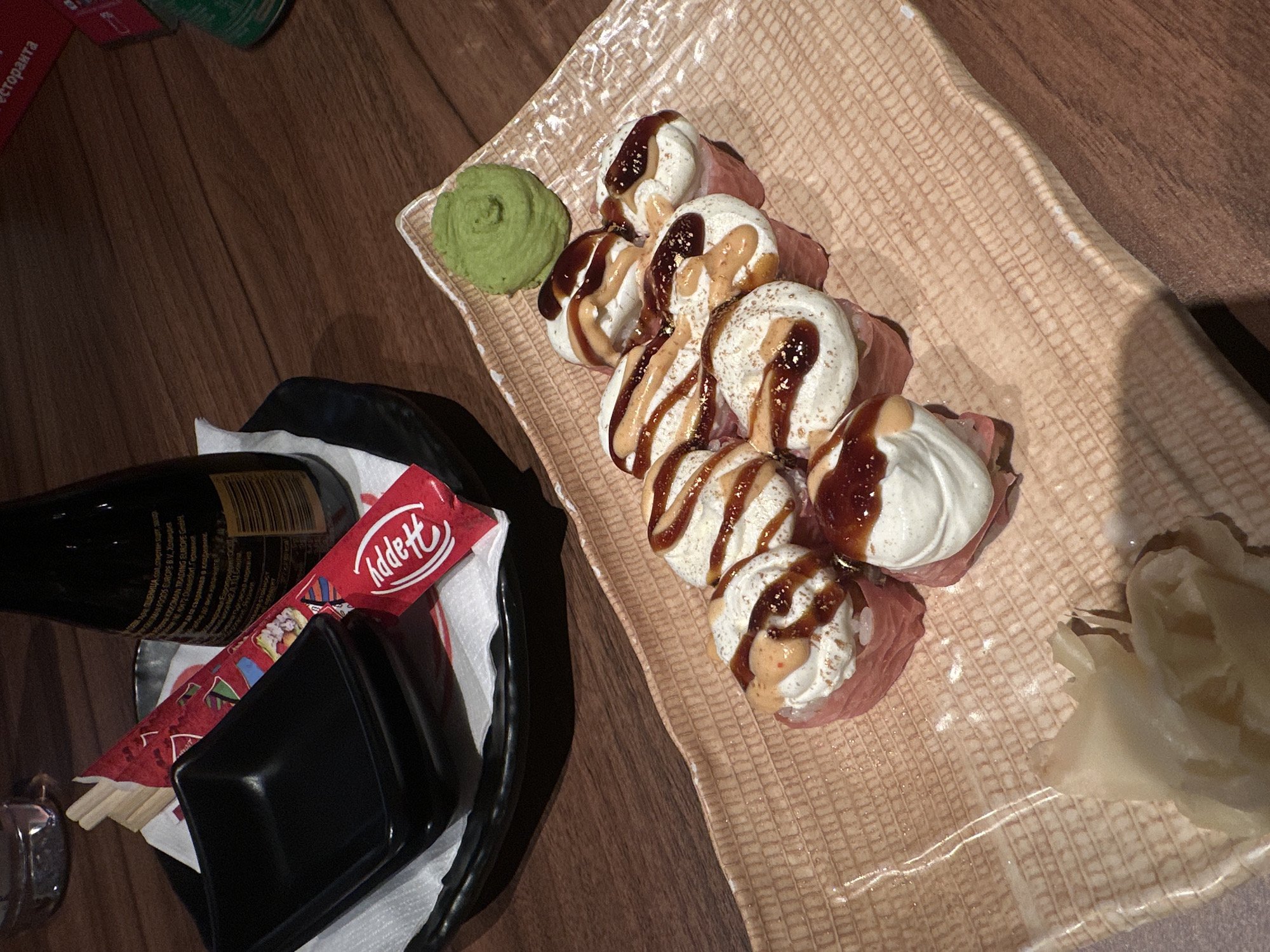 A top-down, angled shot captures a meal, likely sushi or a similar rolled dish, served on a dark wooden table. The main focus is a rectangular, textured beige plate holding several rolls. Each roll features a core wrapped in what appears to be thin slices of meat or fish, topped generously with a white, creamy substance, and drizzled with two distinct brown sauces – one darker and glossy, the other lighter and opaque, with some showing subtle gold flecks.
Accompanying the rolls on the plate are a vibrant green swirl of wasabi paste to the left and several pale, translucent slices of pickled ginger to the right.
In the bottom left corner of the frame, a black bottle, likely containing soy sauce, is visible with its label facing upwards. The label has text in Bulgarian, including "ТЪРГОВСКА МАРКА" (TRADEMARK), "ПШЕНИЦА" (WHEAT), "500DS EUROPE B.V.", "ХОЛАНДИЯ" (NETHERLANDS), "ГЕРМАНИЯ" (GERMANY), and an instruction "СЪХРАНЯВАЙТЕ НА ХЛАДНО И СУХО МЯСТО" (STORE IN A COOL AND DRY PLACE). Part of the word "РЕСТОРАНТА" (RESTAURANT) is also visible. Alongside the bottle are a pair of chopsticks in a red paper wrapper that reads "Addett" and partially "Happy", and a small black, square dipping bowl. Part of a green can is also partially visible in the upper left.
The lighting is soft and warm, suggesting an indoor setting, possibly an evening meal in a restaurant or at home.
