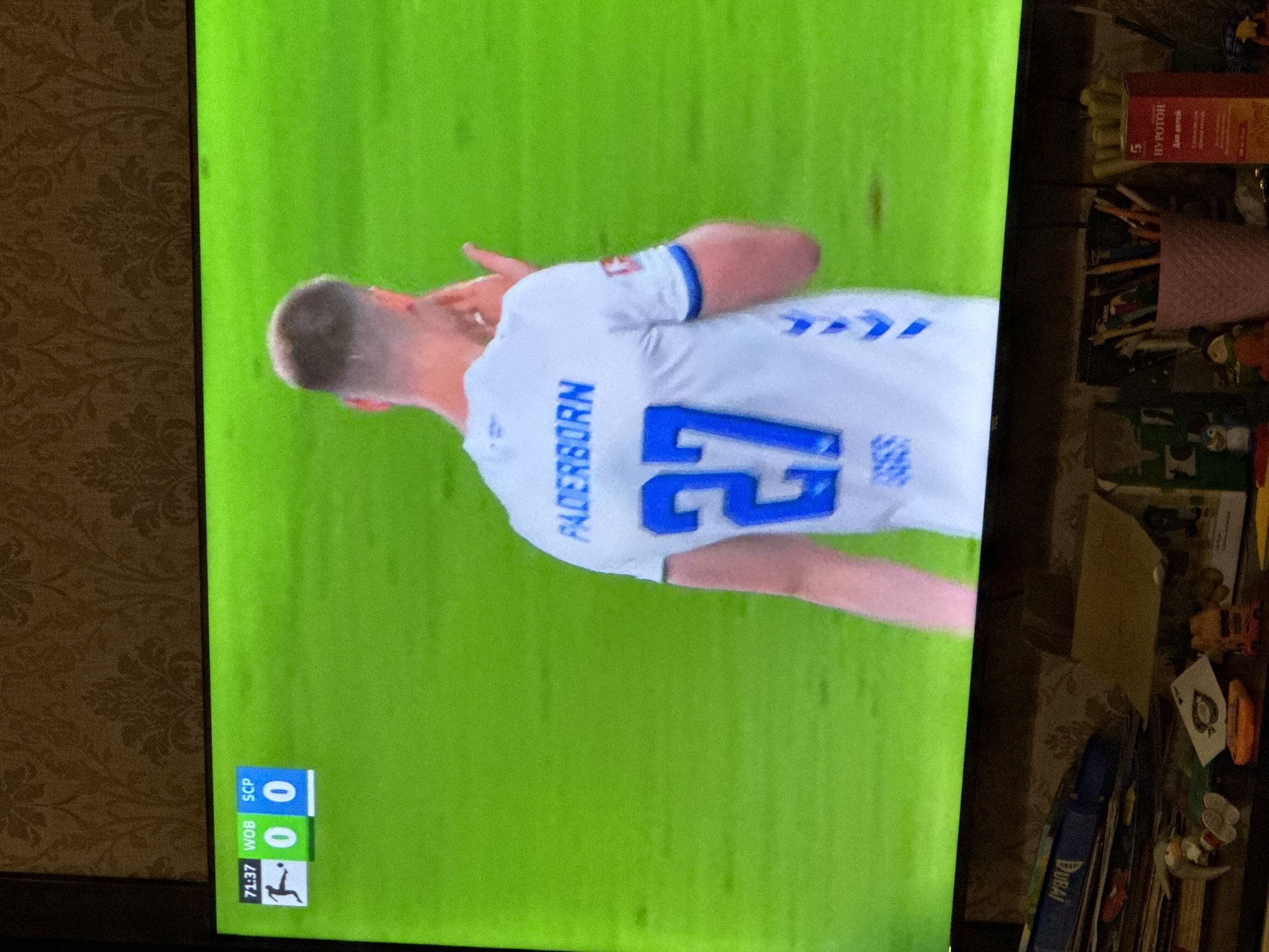 The image shows a television screen displaying a soccer game. The score is WOB 0, SCP 0, and the time is 71:37. A player wearing a white jersey with the name "PADERBORN" and the number "27" is visible. The background appears to be a green soccer field. The TV is placed on a surface with assorted objects, including a box labeled "НУРОТОН" in Russian, stationery, papers, and small figurines. The image was likely taken indoors, with a patterned wallpaper visible in the background. No evidence indicates that the image was taken in Yerevan, Armenia.