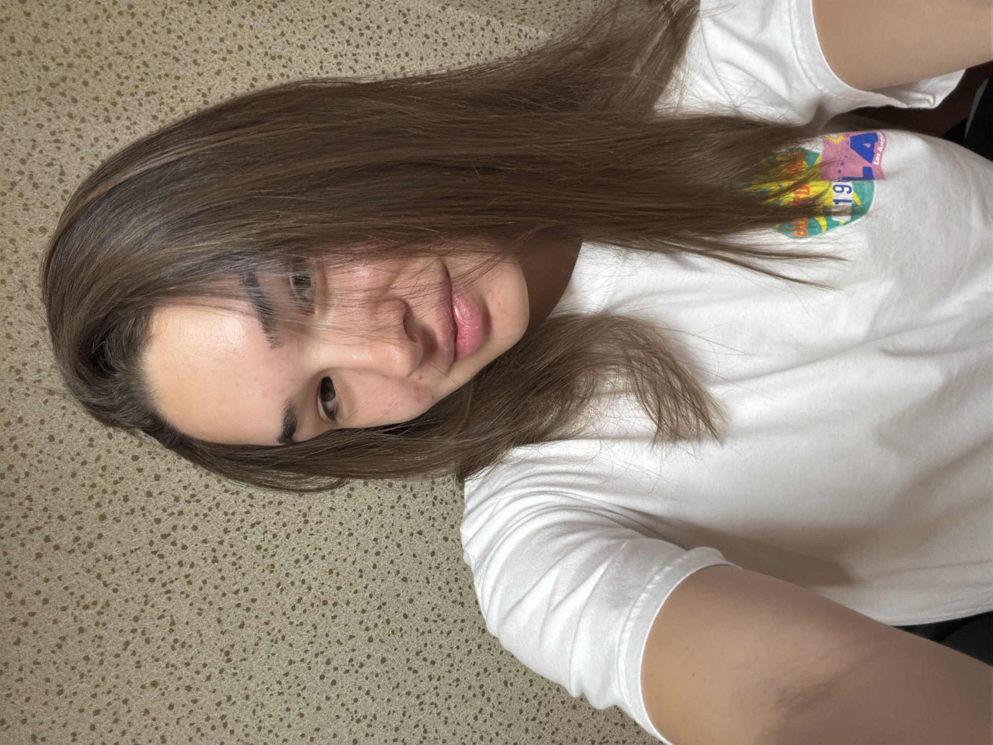 The media file presents a close-up, diagonal selfie of a young person with long, straight brown hair. The subject is lying down, with their head positioned towards the upper left of the frame, and their body extending diagonally across the image. They have a neutral to slightly relaxed expression, with one eye partially obscured by hair and the other looking towards the camera. A faint hint of a smile is visible on their lips.
The person is wearing a white short-sleeved t-shirt. On the chest area, a vibrant, colorful, circular graphic is visible, partially covered by hair. This graphic features a mix of pink, blue, yellow, and green tones, and includes fragments of text, notably the number "19" and what appears to be "LA" or "Los Angeles."
The background is a textured surface, possibly a beige or light brown carpet or rug, characterized by a fine, speckled pattern. The setting is indoors, and there are no discernible details to indicate the time of day or weather. The image was taken in Sofia, Bulgaria, according to the provided context, although no specific landmarks or cues within the photo itself confirm this location. The overall atmosphere conveyed is casual and candid.