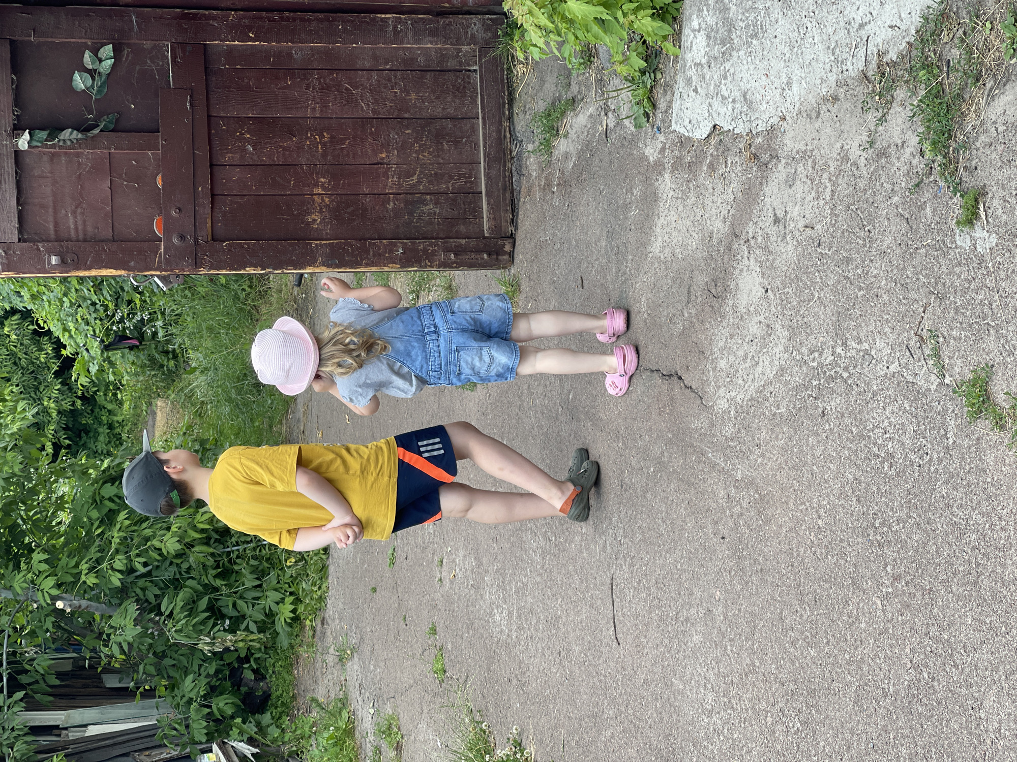 In an outdoor setting, likely a backyard, two children stand on a paved path. A boy, wearing a yellow t-shirt, dark shorts with orange accents, a dark baseball cap, and green clogs, stands facing away from the camera with his hands clasped behind his back. To his right and slightly in front, a young girl, facing away from the camera, wears a light pink hat, a gray t-shirt, denim overalls, and pink Croc-style sandals. She appears to be looking at a weathered, dark brown wooden door or gate on the right side of the frame. The environment is lush with green foliage and trees in the background. The paved path shows some cracks and is bordered by grass and dirt.