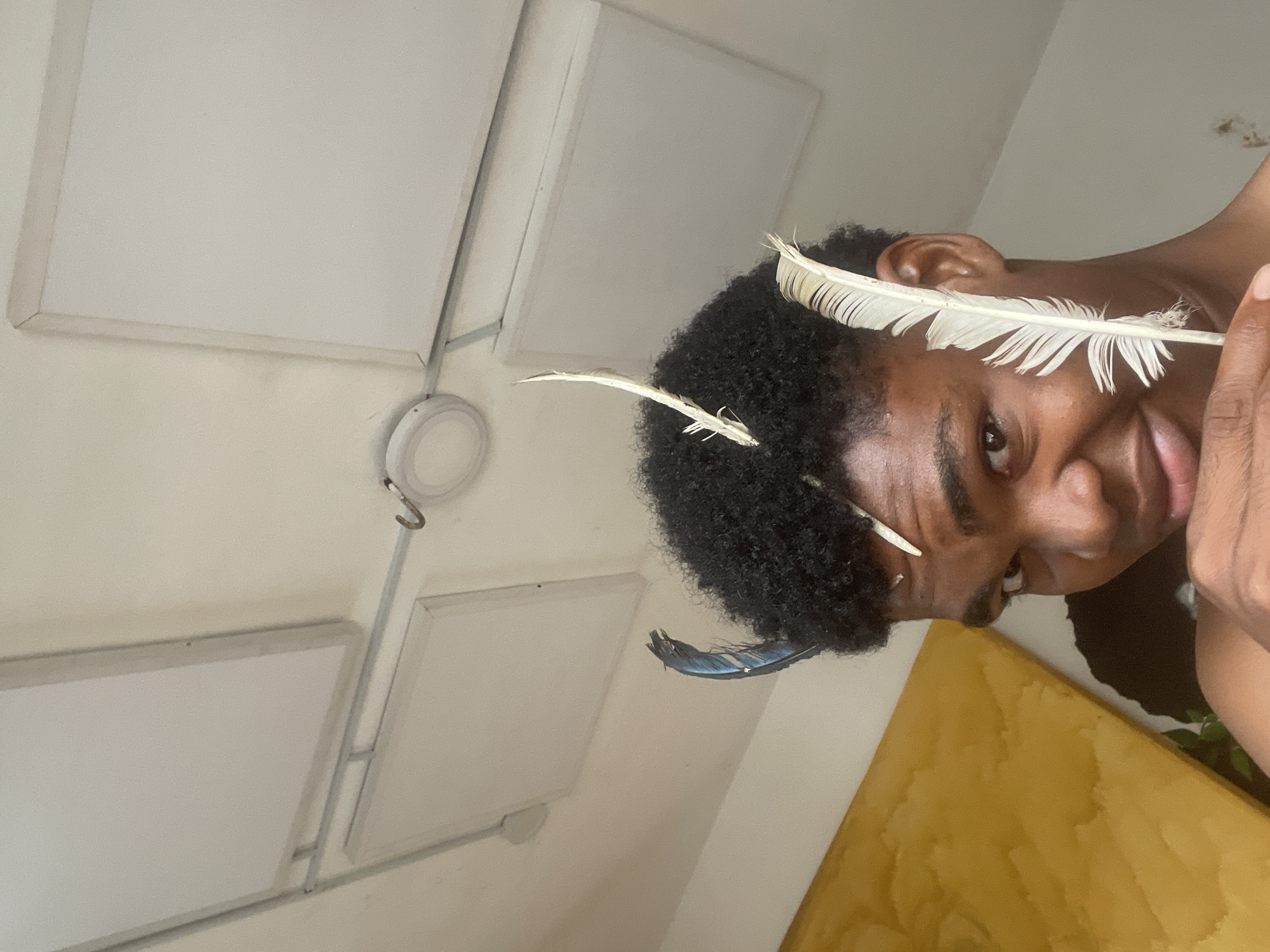 In the city of Ado, Nigeria, an individual with dark, curly hair is looking directly at the camera. They are holding a white feather against their face and have other feathers in their hair, one of which appears blue. The individual is shirtless and positioned indoors, with a yellow object visible to the left and a ceiling with square panels and lights in the background.