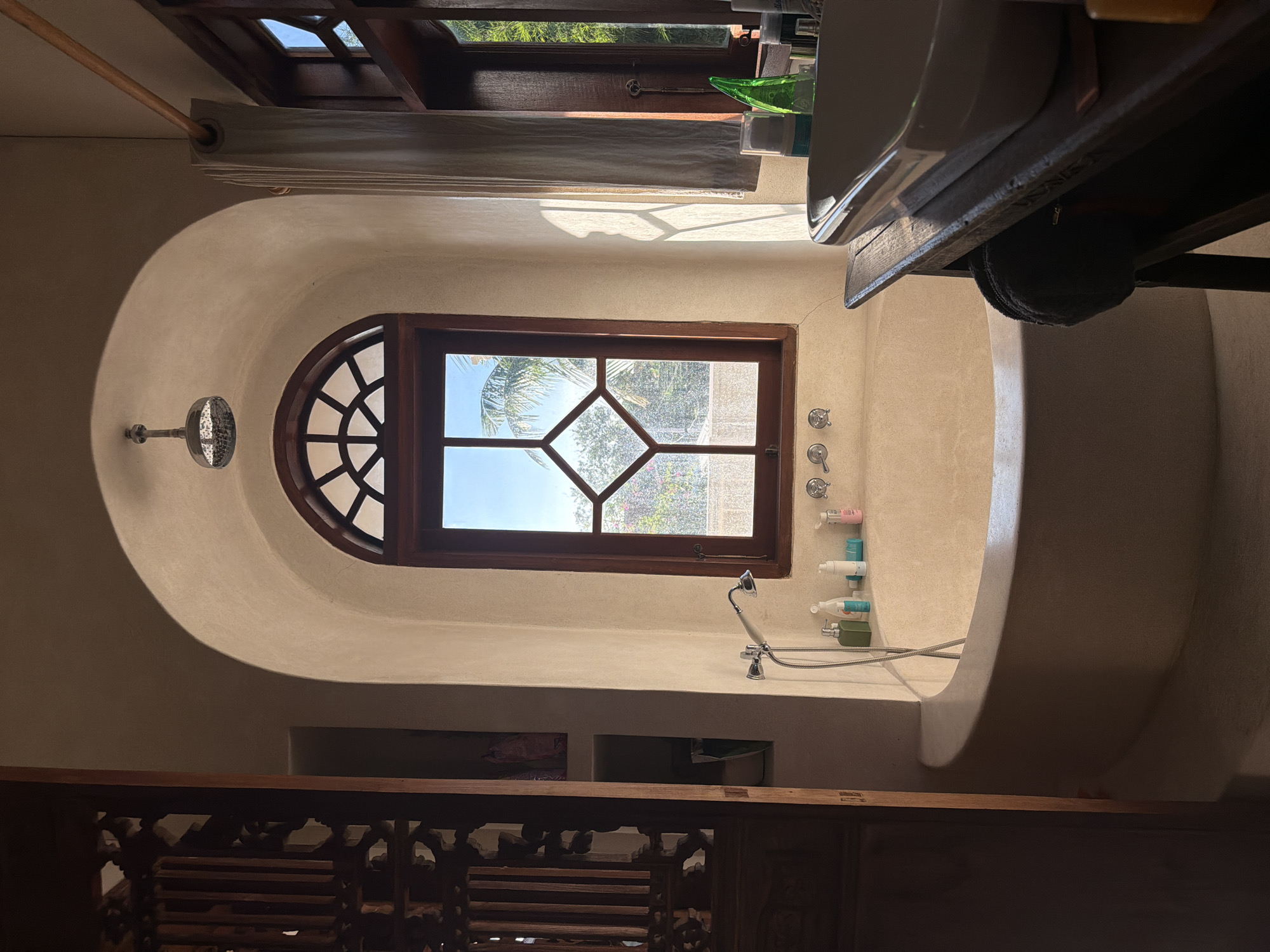 This media file captures the serene interior of a bathroom, likely part of a villa or resort in North Kuta, Indonesia, given the context. The room features organic, curved walls and built-in fixtures, giving it a unique, earthy aesthetic.
The central focal point is a large, arch-shaped wooden window that allows ample natural light to flood the space. The top section of the window has a radial, fan-like grid, while the lower section features a diamond-patterned grid. Beyond the window, lush green foliage and what appears to be part of a palm tree are visible, suggesting a tropical outdoor environment. Sunlight streams through, casting distinct shadows on the curved walls.
To the left of the main window, a fixed showerhead extends from the wall. On the right, a handheld showerhead is mounted near a built-in, curved structure that could serve as a deep bathtub or a shower bench. Several bottles of toiletries are neatly arranged on a ledge alongside this structure.
At the top right of the frame, a white sink basin is visible, resting on a dark wooden vanity unit. Above this area, another window or skylight is partially visible, with a wooden framework and light-colored curtains or blinds.
In the foreground at the bottom, an intricately carved wooden railing or balustrade adds a decorative touch, possibly separating the bathroom from another area or serving as a safety barrier for a step-down. The overall impression is one of natural light, tranquil design, and an integration with the tropical surroundings, characteristic of a daytime scene. No people are present in the image. There is no visible text.