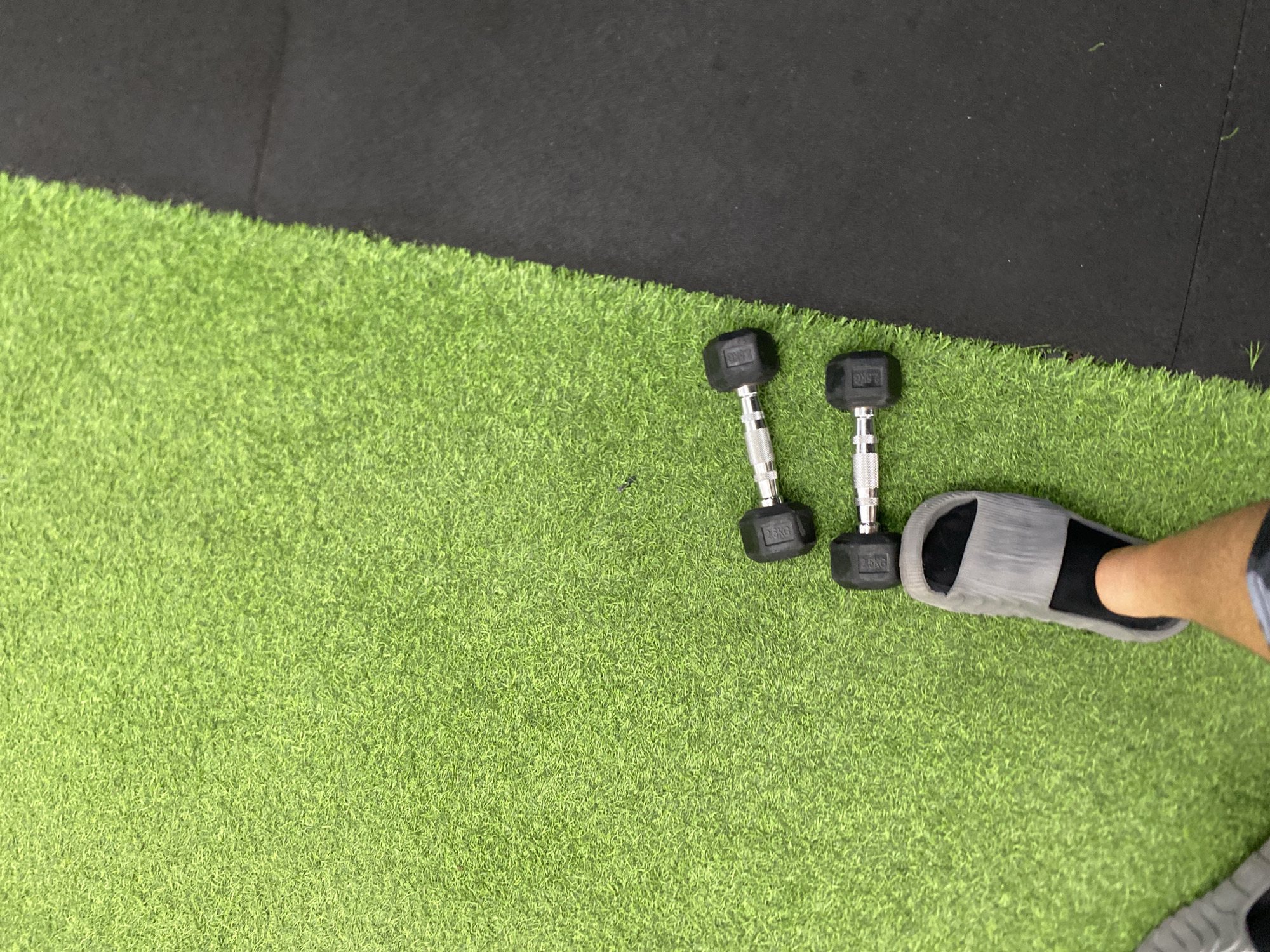 The image captures a top-down view of a gym floor. The floor is divided into two sections: artificial green turf on the left and black rubber matting on the right. Two 2.5kg dumbbells are placed on the green turf. A person wearing gray slides and black socks is standing on the turf, with one leg and foot partially visible. The scene is likely indoors, given the lighting and gym equipment. There is no specific information about the city of North Kuta, Indonesia, in the image.