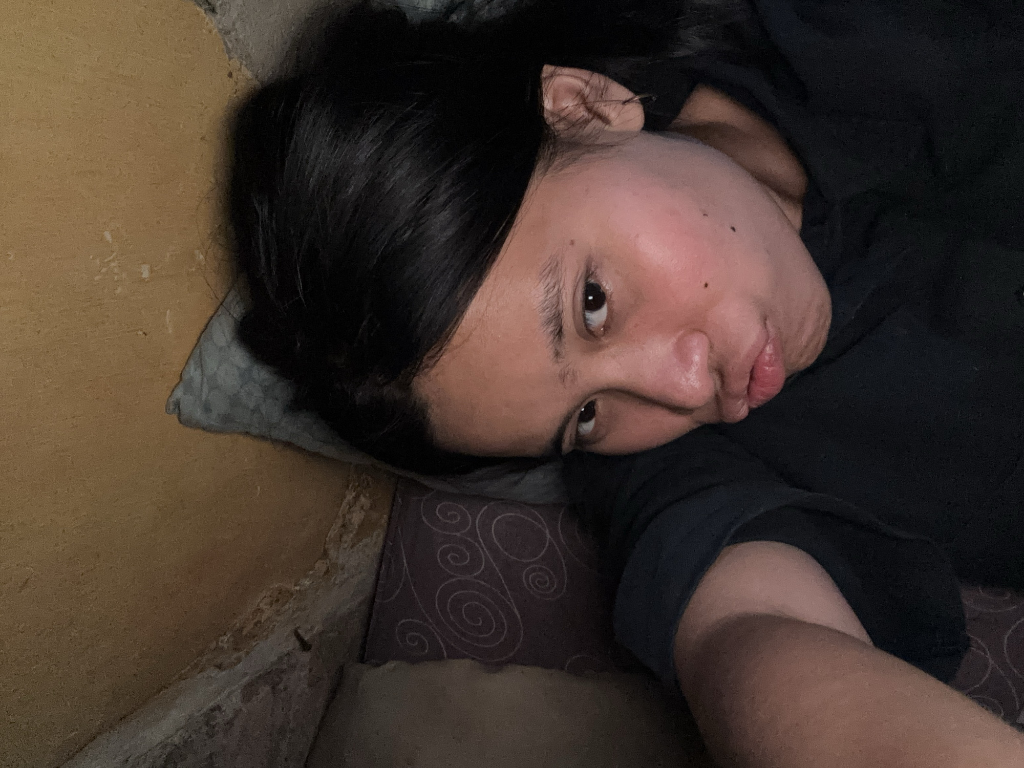 The image shows a person lying on their side, resting their head on a pillow. The person has dark hair and is wearing a dark shirt. Their face is turned towards the camera, with their eyes open and looking slightly to the side. Their lips are slightly parted. The background appears to be a wall with a textured, yellow surface on the left and a darker, possibly tiled or patterned surface on the right. A pillow with a swirling pattern is visible beneath the person's head. The lighting suggests an indoor setting, possibly at night or in a dimly lit room. The overall mood conveyed by the image is one of repose or perhaps slight weariness. There is no visible text in the image. The context provided is East Jakarta, Indonesia.