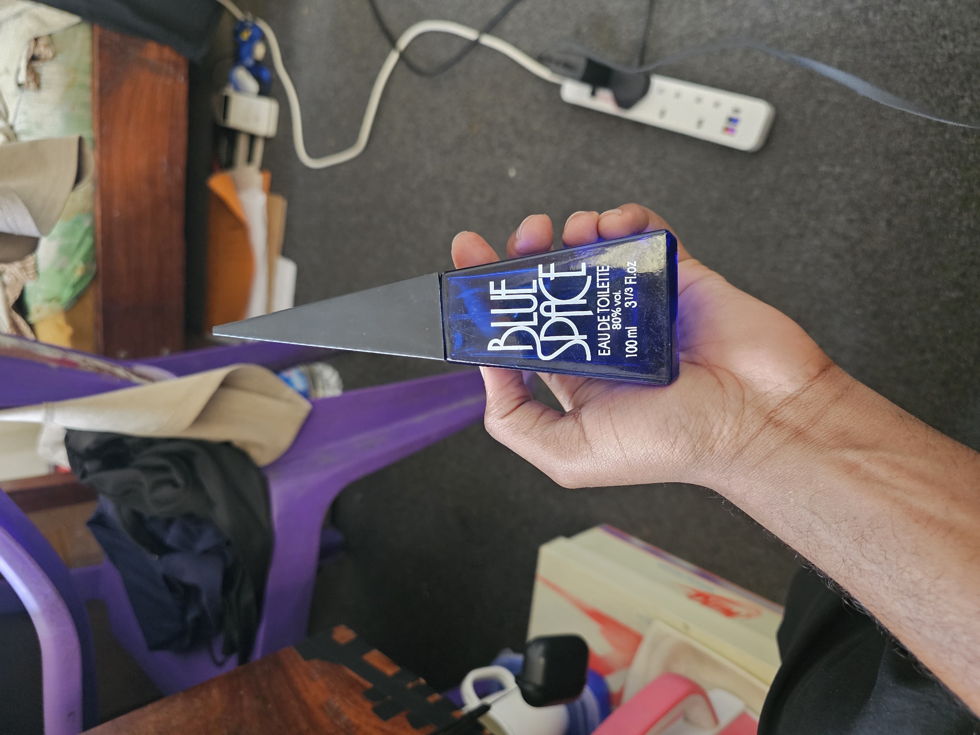 A person's hand holds a blue glass bottle of "BLUE SPACE" eau de toilette. The bottle has a black, triangular cap. The text on the bottle reads "BLUE SPACE EAU DE TOILETTE 80% vol. 100 ml 31/3 Fl.oz". The background is a gray carpeted floor. To the left of the bottle, a white ceramic mug and a black object are visible on a yellow and white patterned surface, possibly a box. Behind this, a purple plastic chair with a black cloth draped over it is present. In the further background, a wooden surface and a power strip with a white cord can be seen. The scene is indoors and appears to be a domestic setting, possibly in Dar es Salaam, Tanzania, given the presence of typical household items.