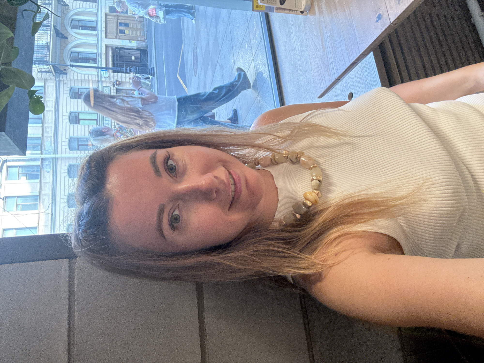 A selfie-style portrait shows a woman indoors, partially obscuring a large window that reflects a city street scene. The woman, with long, light brown hair and green eyes, smiles faintly at the camera. She wears a light-colored, possibly cream or white, ribbed top and a necklace made of large, irregularly shaped, light-colored beads. Her arm is partially visible on the right. She appears relaxed.
Behind her, through the window, an urban environment is reflected. Detailed historic-looking buildings with arched windows and decorative facades line a street. The reflection shows people walking on a paved sidewalk or street. One person with long, lighter hair and blue jeans is prominently reflected, along with another person further back. A plant with green leaves is visible on the far left, also reflected in the window.
Indoors, a dark, textured surface, possibly a table, is visible to the woman's right. A dark grey upholstered surface is seen below and to her left.
The scene takes place during the daytime, indicated by the natural light. The overall mood is calm and casual. Visible text in the window's reflection includes "LUCKY KITCHEN" and "HAND SANITISER," possibly from a sticker or sign inside the establishment where the photo was taken. The architectural style suggests a European city setting.