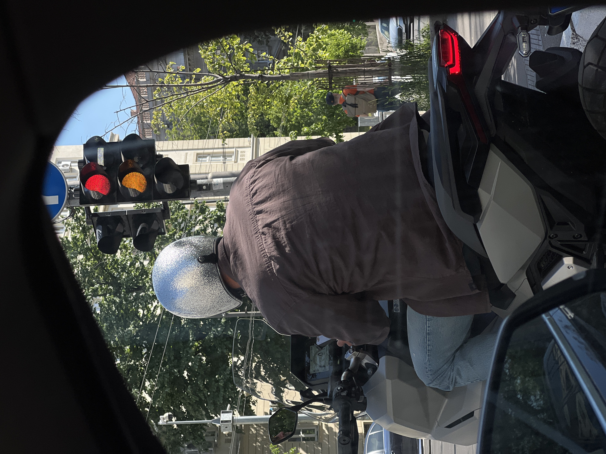 The image, taken from inside a car, shows a person on a scooter stopped at a traffic light in Belgrade, Serbia. The traffic light is displaying both a red and yellow light. The person on the scooter wears a silver helmet, a brown jacket, and blue jeans. The scooter is light gray and black. Through the car window, buildings and trees typical of Belgrade can be seen. The presence of a blue traffic sign indicates the direction of traffic flow. The scene appears to be a typical urban street setting.