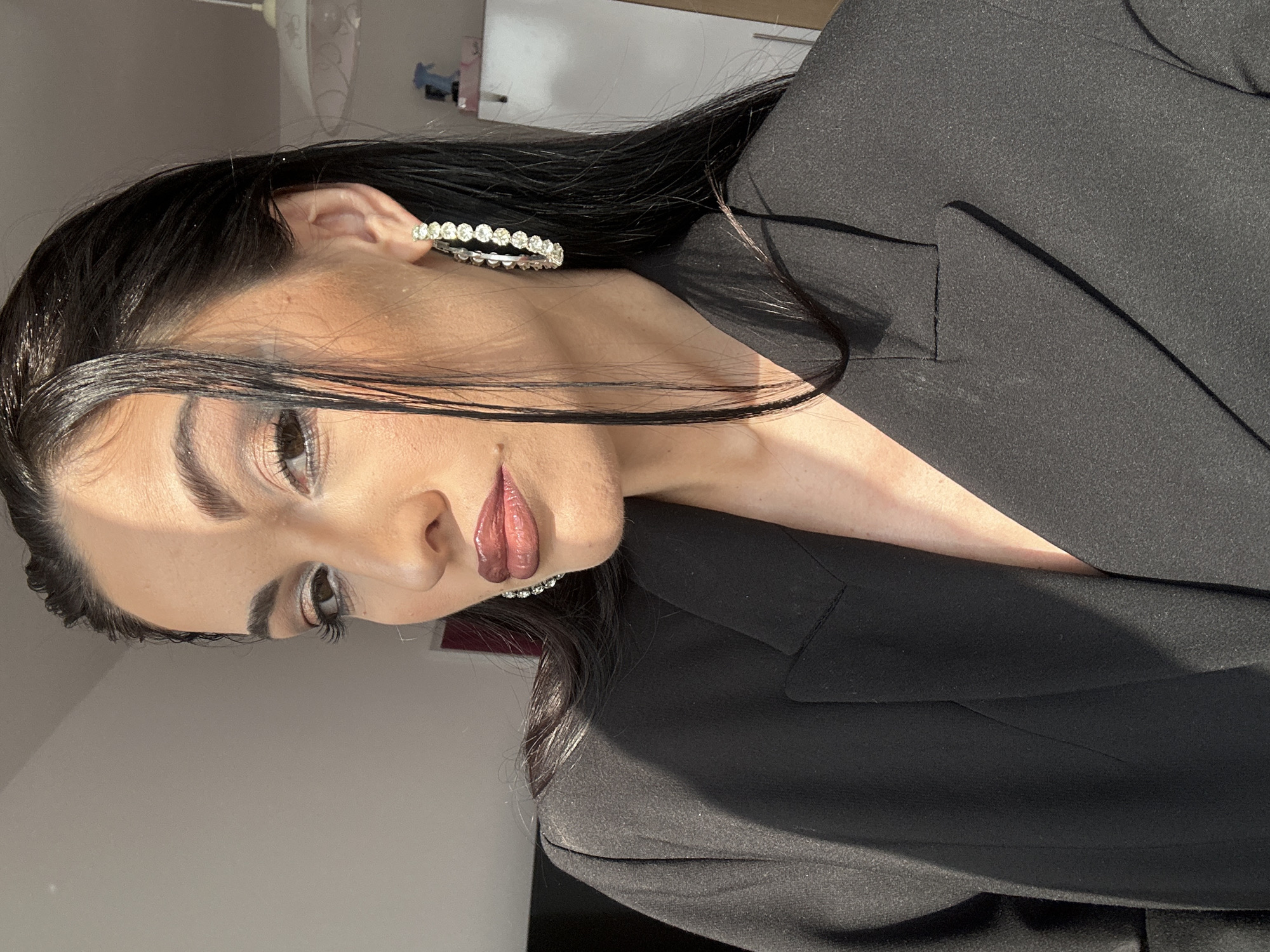 The image displays a close-up portrait of a woman, angled sideways. Her head is tilted, and she looks slightly away from the camera. She has long, dark, straight hair, which frames her face and falls over her shoulders.
She is wearing a black blazer, visible on her upper body and shoulder. A prominent feature is a large, circular hoop earring on her visible ear, studded with multiple small, sparkling stones, likely diamonds or crystals. Her makeup includes neatly groomed eyebrows, extended black eyeliner with a slight wing, and glossy, dark pink or plum-toned lipstick. Her eyes are dark brown.
The scene is illuminated by strong, natural sunlight, casting bright highlights on her face and hair, particularly on the left side, and creating distinct shadows. The background is a plain, light-colored wall, suggesting an indoor setting. Hints of furniture or fixtures are barely discernible in the top background. The strong lighting suggests it is daytime. There is no visible activity beyond posing. No text is visible in the media file.