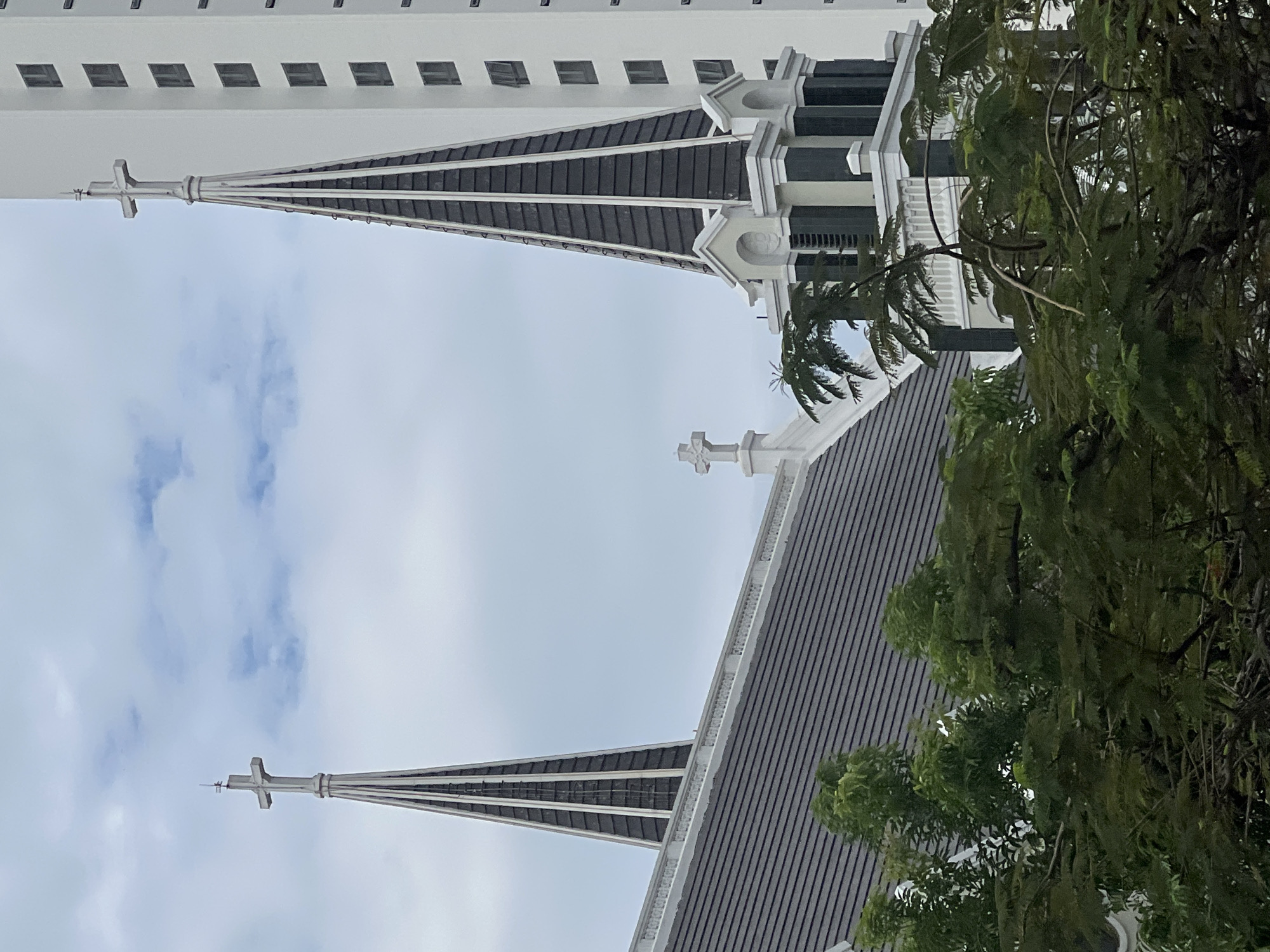 The image captures a multi-layered architectural scene in Bac Nha Trang Ward, Vietnam, under a bright, overcast sky. The primary subjects are two ornate spires, likely belonging to a church, and a tall, modern building.
In the foreground and mid-ground, two distinct church spires rise towards the sky. The more prominent spire features a slender, dark-tiled cone culminating in a light-colored cross at its apex, with additional decorative white elements and finials near its base. Further back, another similar spire with a cross is partially visible. A substantial section of a dark-tiled roof, adorned with a white balustrade and another cross-shaped ornament, forms part of the church complex in the lower portion of the image.
To the right, lush green foliage from large trees, possibly including palm fronds, partially obscures parts of the church structures, adding a natural element to the urban landscape. In the background, a tall, white building with multiple rows of dark, rectangular windows stands, presenting a stark, modern contrast to the traditional architecture of the church.
The sky is bright and cloudy, suggesting daytime conditions without direct sunlight. There are no people visible, and no specific activities are taking place. The image is devoid of any discernible text. It presents a serene juxtaposition of religious and contemporary architecture.
