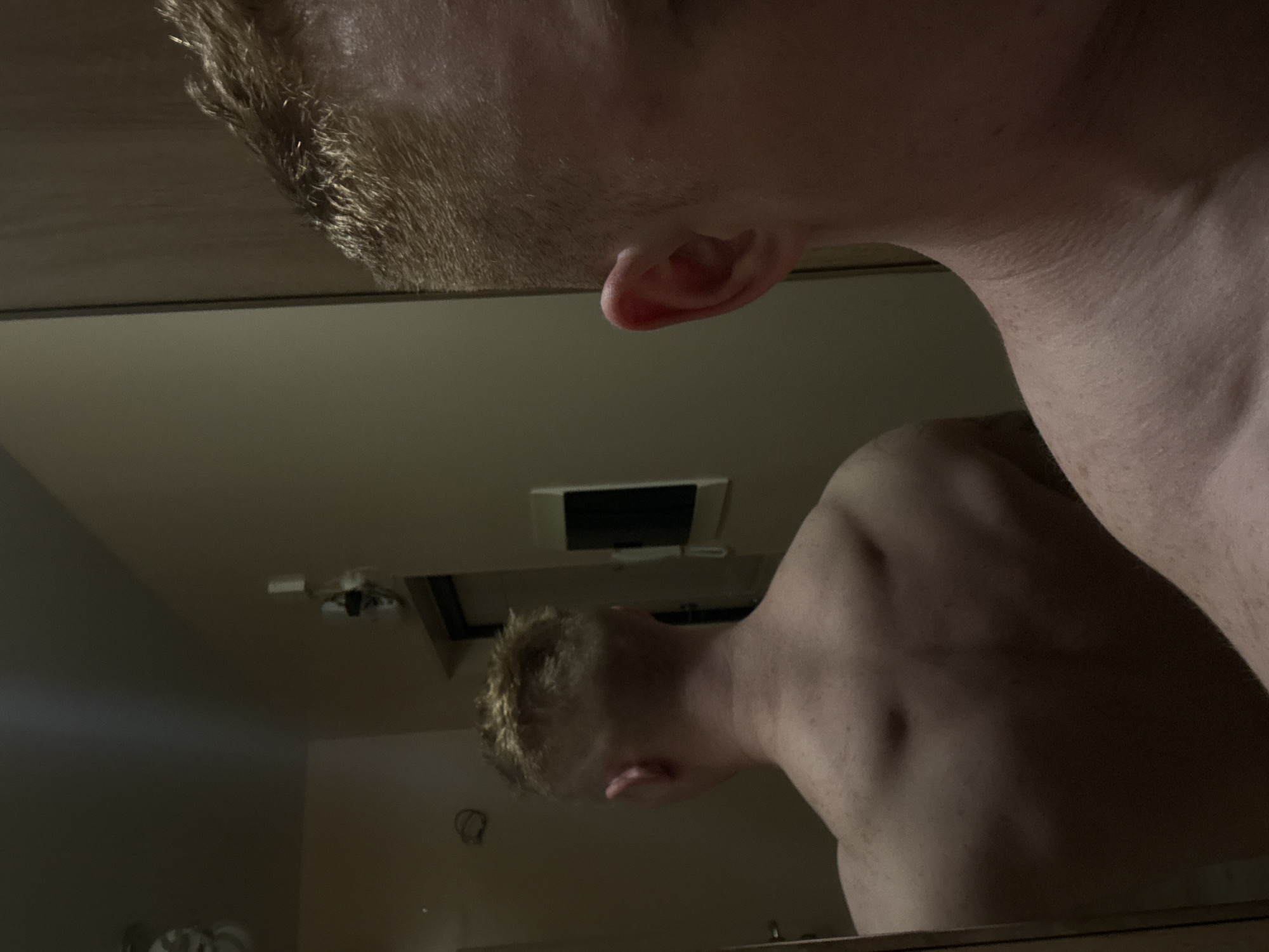 A pale-skinned person with short, light-colored hair is looking at themselves in a mirror. The person is shirtless, and their ear is visible. The mirror reflects the ceiling, which has a vent and a light fixture. The location is likely indoors in Chaikovsky, Russia, but there is insufficient detail to confirm.
