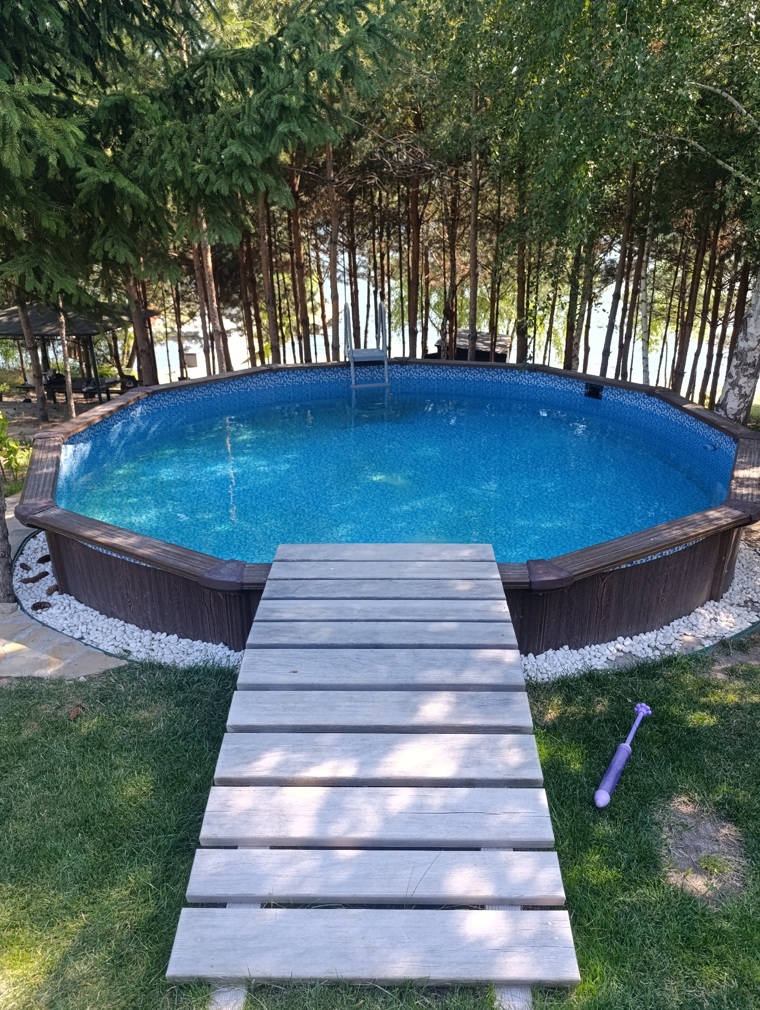 The image shows an above-ground swimming pool surrounded by a wooden deck and lush green grass. Tall trees, predominantly pine and birch, frame the scene, with a body of water visible in the background. A wooden walkway leads to the pool. A purple water gun is lying on the grass to the right of the walkway. The overall impression is of a recreational area in a natural setting. The location is identified as Bulatselivka, Ukraine.