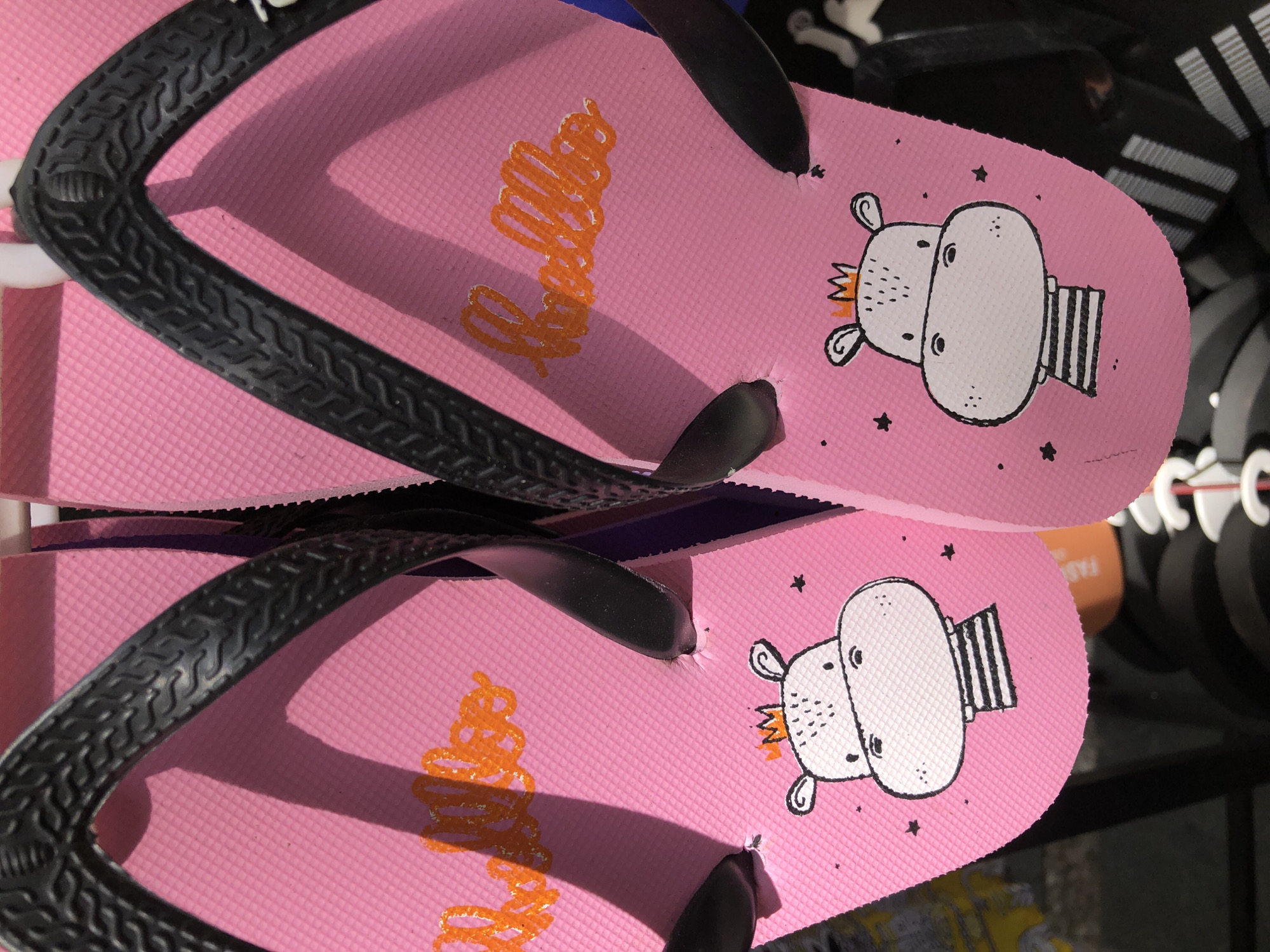 This media file shows a pair of pink flip-flops with black straps. The flip-flops have a graphic design featuring a cartoonish hippopotamus wearing a crown and a striped shirt. Small black stars are also scattered around the hippopotamus. The word "Hello" is written in orange script on the flip-flops. The flip-flops appear to be new and are displayed in a retail setting. The background is dark, with some white hooks and other footwear visible. The lighting suggests an indoor environment, possibly a store. The overall mood is lighthearted and playful due to the cartoon design.