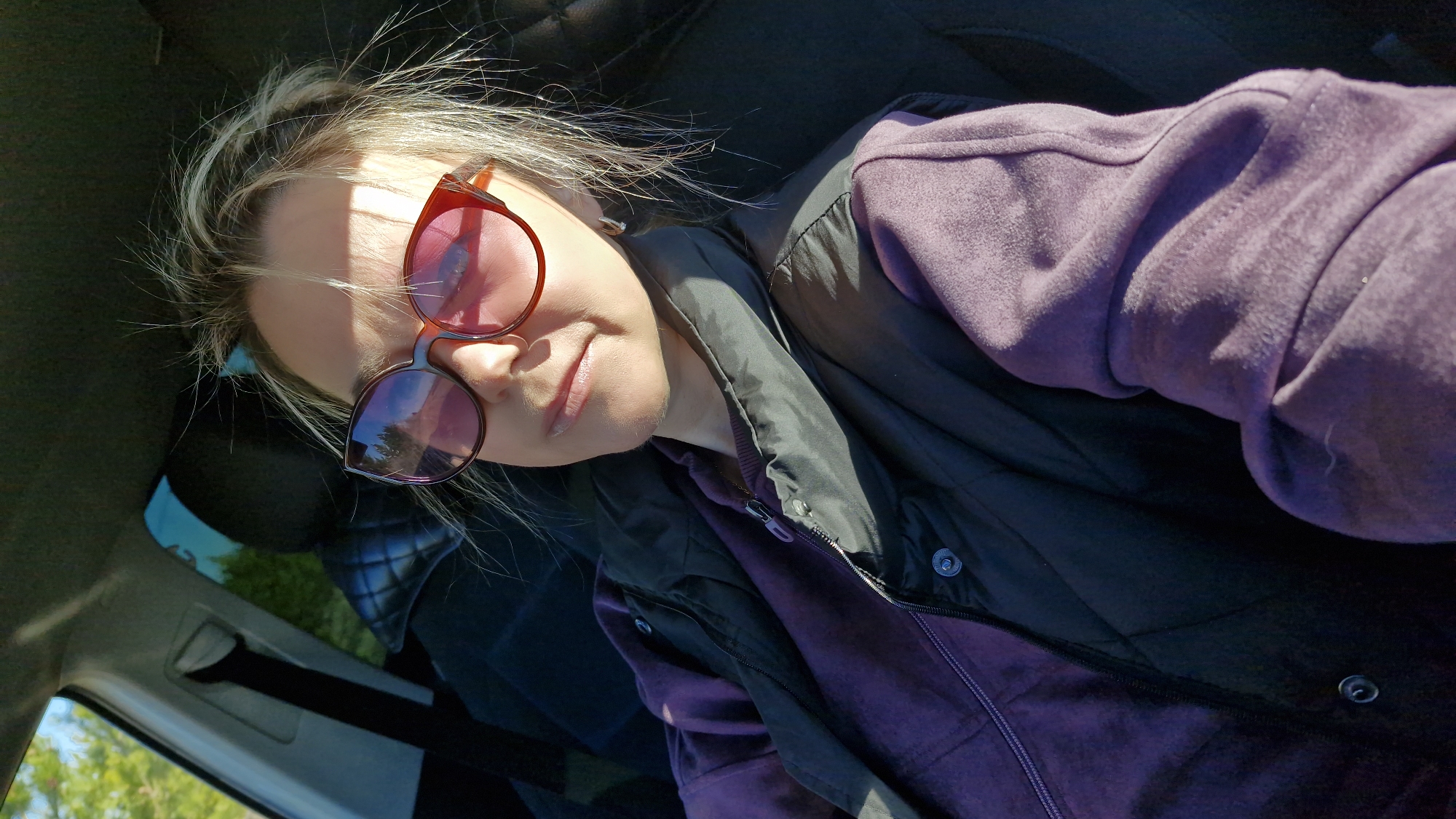 A person, likely female, is captured in a sunlit selfie from a high angle, appearing to be inside a car. She has blonde hair and is wearing large, reddish-brown framed sunglasses with pink-tinted lenses, which reflect trees and a bright sky. A distinct shadow from the sunglasses or an overhead object falls across her forehead and nose due to strong direct sunlight. She wears a textured purple long-sleeved top, possibly fleece or velour, underneath a dark, likely black or deep purple, quilted vest or jacket with metal snap buttons and a zipper. Her expression is a subtle, relaxed smile.
The background reveals parts of the car's dark interior, including what appears to be a seat with a quilted pattern and a seatbelt mechanism. Outside the car window, hints of green foliage and a bright blue sky suggest an outdoor, daytime setting. The image conveys a casual, bright moment, possibly while traveling or parked in Otchor, Russia. No visible text is present.