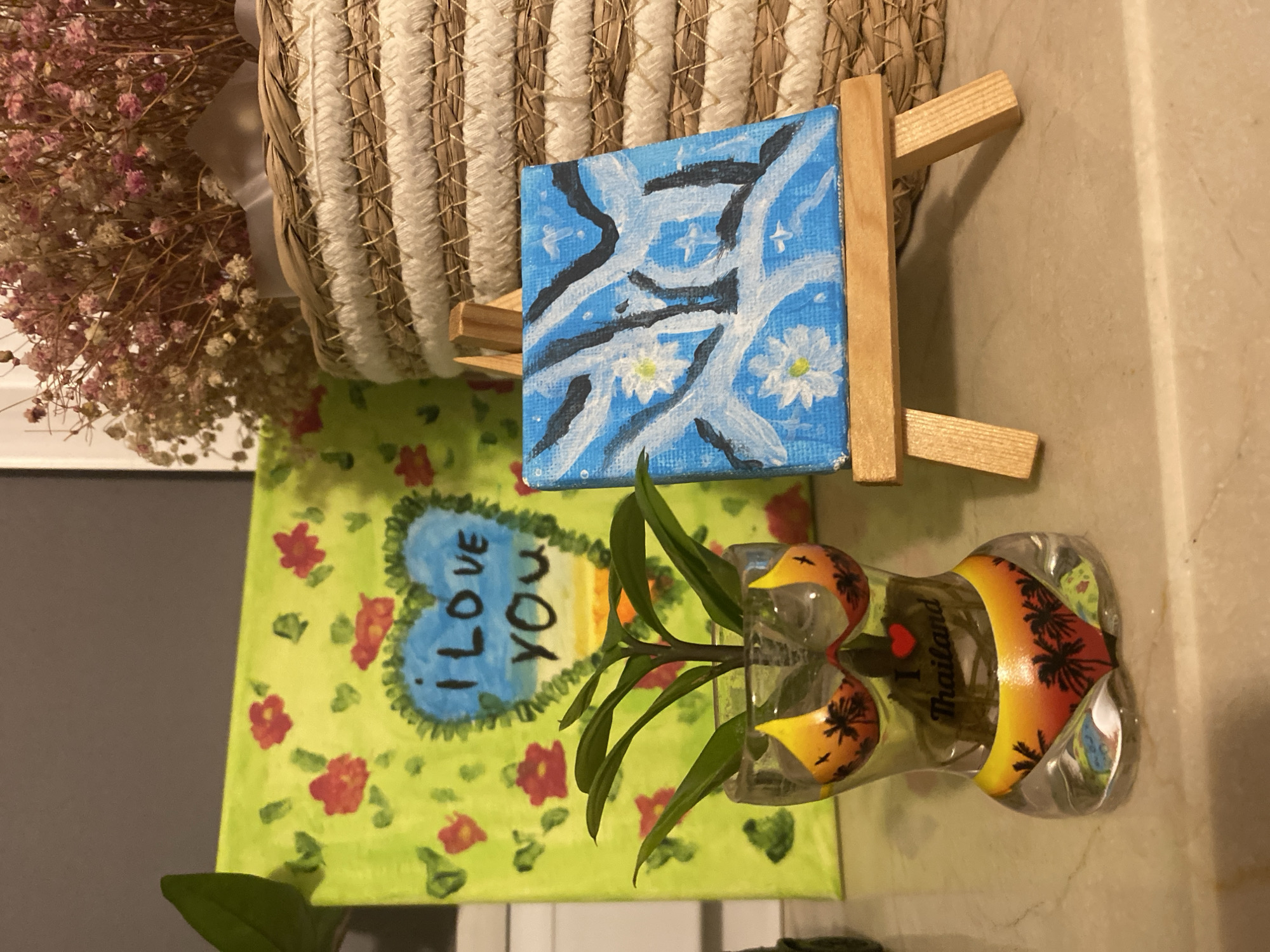 The image depicts a still life arrangement on a marble-like surface. In the foreground, to the left, is a clear glass planter shaped like a female torso, filled with water and a small plant with green leaves. The planter is decorated with a bikini pattern featuring a sunset scene with palm trees and the text "I <3 Thailand." To the right of the planter, a small canvas painting of white flowers against a blue background is displayed on a wooden easel. Behind these items, a larger canvas painting with a lime green background, red flowers, and a blue heart with the text "I LOVE YOU" is leaning against an unseen surface. To the right of the paintings, a woven basket filled with dried pink and white flowers adds to the composition. The overall lighting suggests an indoor setting. The location in the city of Łódź, Poland, cannot be determined from the visual information provided in the image.