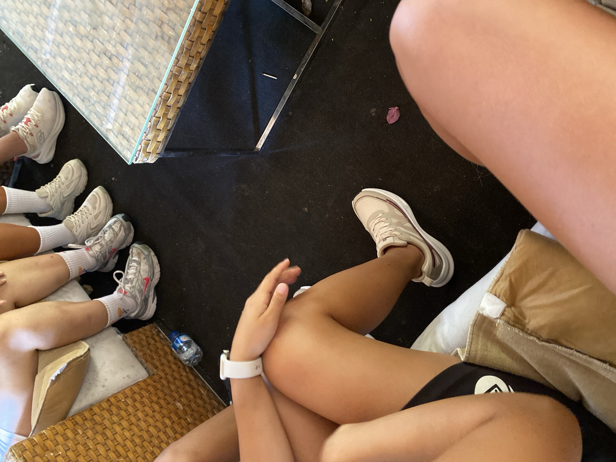 The image depicts a close-up view of several individuals' legs and feet, suggesting they are seated or resting on a low surface. The setting appears to be outdoors or in a well-lit indoor space with a dark carpeted floor.
In the foreground, a person's bare legs are visible, one bent at the knee and the other extended. They are wearing beige sneakers with white soles and burgundy accents. A white watch is worn on the left wrist. To the left of these legs, another person is partially visible, wearing black athletic shorts and a beige-brown blanket or cushion.
Further back, several other individuals are seated, their legs also visible. They are all wearing white athletic socks and various styles of athletic shoes. Some are wearing white or light-colored sneakers, while others have shoes with more prominent detailing and branding, such as a pink Nike swoosh. One person is wearing white socks with a colorful, abstract pattern.
A clear plastic water bottle with a blue cap is lying on its side on the carpet near the seated individuals. A woven rattan furniture piece, possibly a chair or ottoman, is visible in the upper left quadrant of the image. To the right, a modern coffee table with a glass top and a woven rattan frame is present. A single red flower petal is on the dark carpet in the lower right section of the image, and a thin, light-colored twig or straw is near the table leg.
The lighting suggests daytime. The image does not provide any explicit visual cues to pinpoint its location to Aksu, Turkey.
