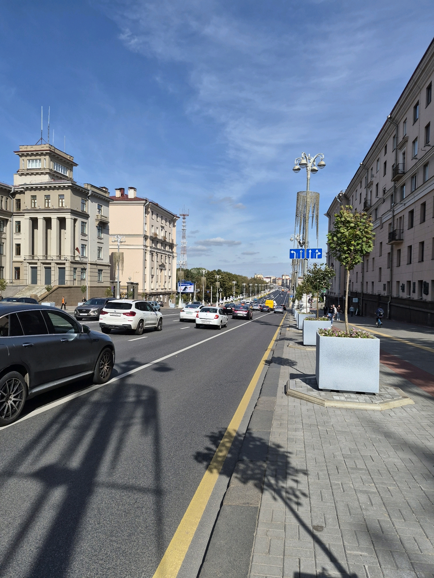 The image depicts a street scene in Minsk, Belarus, on a sunny day. The perspective is from a sidewalk looking towards a multi-lane road with moderate traffic. On the left, there are several large buildings with classical architectural elements, including columns and ornate facades. One building has a tower with antennas. On the right side of the road, there is a sidewalk with patterned paving stones, large planter boxes with trees, and a lamppost with multiple lights. The road has a yellow line separating traffic lanes and a white line marking the edge of the lane. Several cars, including SUVs and sedans, are visible on the road. A few pedestrians are seen on the sidewalk and near the buildings. The sky is blue with some scattered white clouds. The overall impression is that of a well-maintained urban environment.