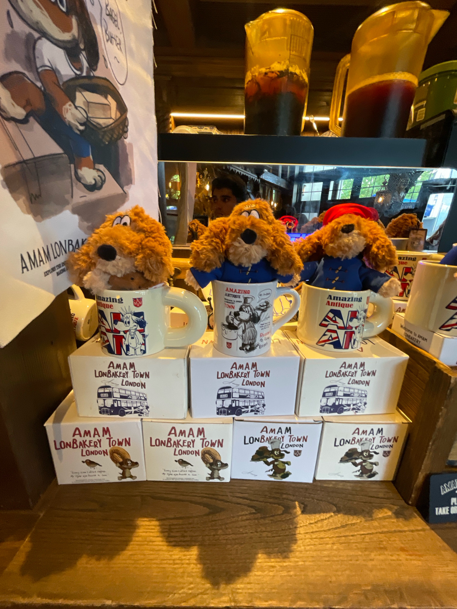 In a Shanghai bakery, three plush toy dogs wearing blue coats sit in mugs on display boxes. The mugs and boxes feature the "AMAM LONBAKERY TOWN LONDON" branding and illustrations of London landmarks like double-decker buses and Union Jacks. Behind the toys, a large fabric banner depicts a cartoon dog holding a basket of goods. On a shelf above, large pitchers filled with dark liquid are visible. The scene is lit by warm, ambient lighting, creating shadows on the wooden surface in the foreground.