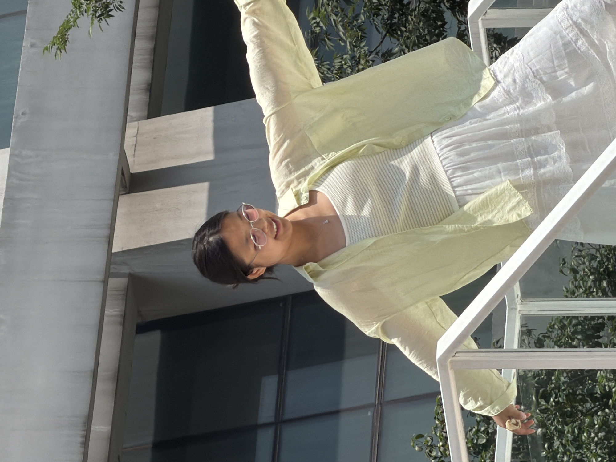 A young Asian woman wearing a light yellow unbuttoned shirt over a white ribbed tank top and a white tiered lace skirt is leaning against a white railing. She is wearing pink-tinted round glasses and a delicate silver necklace with a small pendant. Her right hand is resting on the railing, and her left arm is extended outwards. She is positioned on a white staircase with reflective glass panels. In the background, there is a modern building with concrete architectural elements and tinted glass windows. Green foliage is visible around the railing and the building. The scene appears to be outdoors during the day, with bright lighting and shadows. The location is identified as Yuzhong District, China.