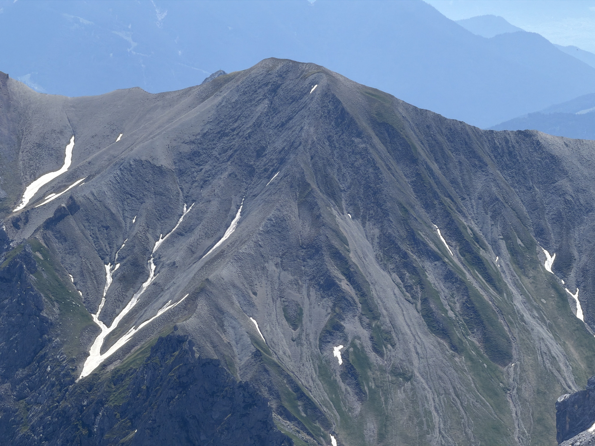 This is a medium shot, eye-level, outdoor photograph of a mountain range in Garmisch-Partenkirchen, Germany. The foreground features a steep, rocky mountain slope with patches of snow and sparse green vegetation. The texture of the mountain is rough and uneven, with visible strata and erosion patterns. Several distinct snow patches, some elongated and others more amorphous, are scattered across the grey rock face. In the background, successive layers of lighter blue mountains recede into the distance, suggesting a vast and expansive landscape under a clear, pale blue sky. The lighting appears to be natural daylight, casting subtle shadows that highlight the topography of the mountains. There are no people or man-made objects visible in the scene.