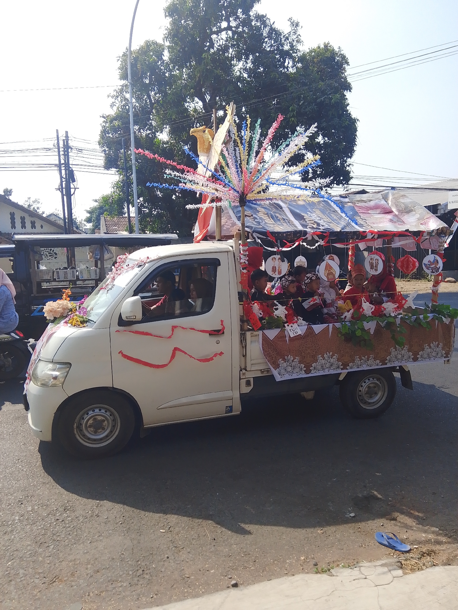 A white pick-up truck is decorated and carrying children in a procession on a street in Kalinyamatan, Indonesia. The truck's bed is adorned with red and white fabric, green foliage, and decorative paper cutouts. Several children, some wearing traditional attire and head coverings, are seated within the decorated bed. The truck's front is also decorated with flowers and red ribbons. The scene is set outdoors on a paved road, with trees, buildings, and utility poles visible in the background. The sky is clear and bright.