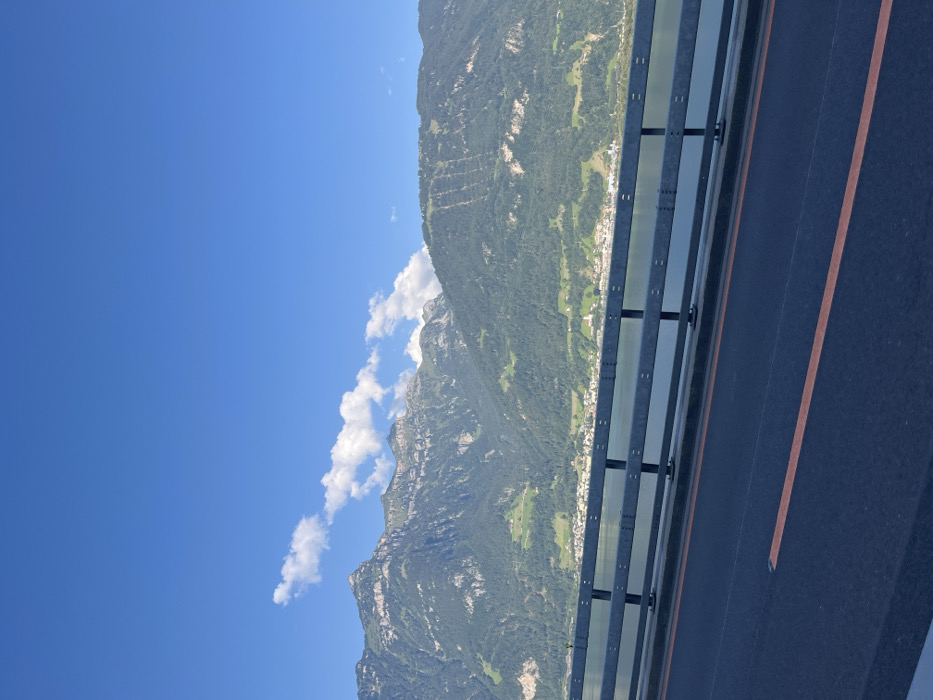 The image depicts a static, wide-angle view from a bridge, likely on the Axenstrasse in the vicinity of Seedorf (UR), Switzerland. The immediate foreground features a section of dark asphalt roadway, angled slightly right, with a single orange lane marking running parallel to the bottom edge. A grey, two-tiered metal guardrail, parallel to the roadway, spans the lower midground, serving as a barrier. Beyond the guardrail, a calm body of water, identified as Lake Lucerne (Urnersee), extends horizontally across the frame. The opposing shoreline, situated in the midground, hosts a cluster of light-colored structures indicative of a small village, nestled at the base of large, dark green, forested mountains. The mountainous terrain dominates the background, displaying varied slopes, some with visible terracing and rocky outcrops. The sky above is clear blue, dotted with scattered white cumulus clouds positioned above the mountain ridge. No human figures or discernible actions are present within the frame.