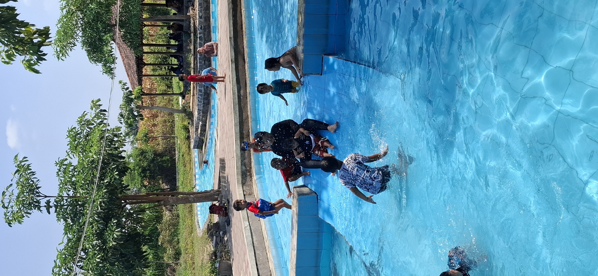 In the city of Unknown, Unknown, on a sunny day, a group of children and adults are enjoying a water park. The scene features a bright blue swimming pool with multiple levels and a slide. Several children are actively playing in the water, splashing and sliding. One child, wearing a blue and red swimsuit, is standing at the edge of a raised platform, appearing to be about to enter the water. Another child, in a patterned shirt and dark shorts, is in the middle of the pool, interacting with the water. An adult, dressed in black, is seated in the pool, holding a young child. Other children are visible in the background, some near the slide and others in different sections of the pool. The surrounding environment includes lush green trees and a covered seating area with people relaxing under it. The ground around the pool is paved with brick-like tiles. The sky is clear and blue, indicating fair weather.